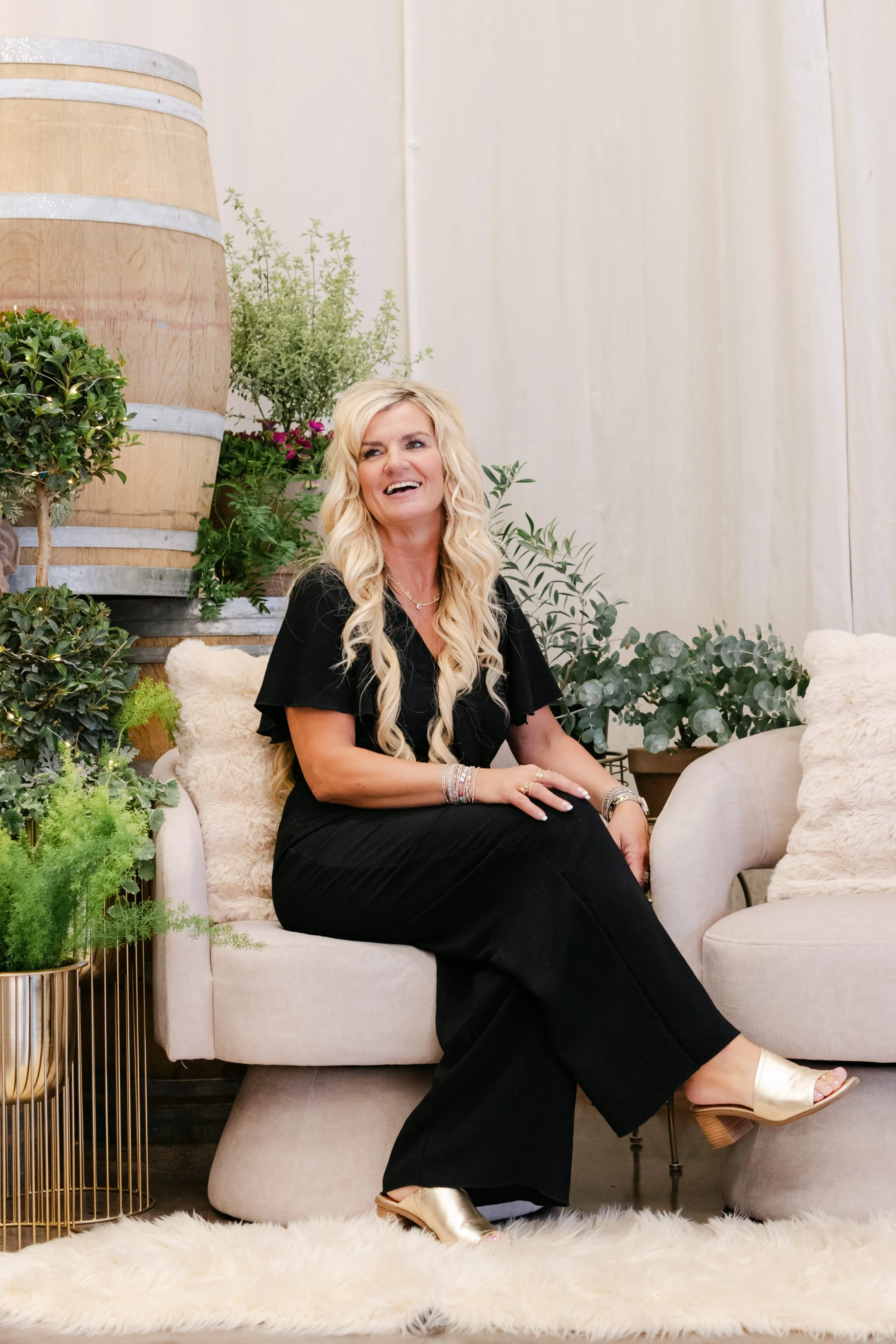 A woman with long blonde hair sitting on a cream-colored chair in a room decorated with green plants and flowers, wearing a black outfit and gold shoes, smiling and looking to her left.