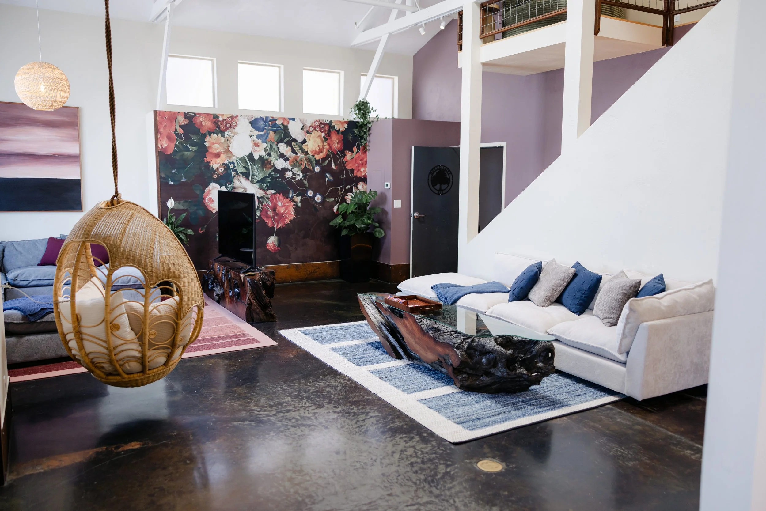 A modern living room with white and purple walls, featuring a floral accent wall and a high ceiling with skylights. The room has various furniture pieces including a white sofa with blue and gray cushions, a wooden table, a rattan hanging chair, and a patterned area rug. There is a large flat-screen TV on a stand, a tall indoor plant, and artwork on the walls.
