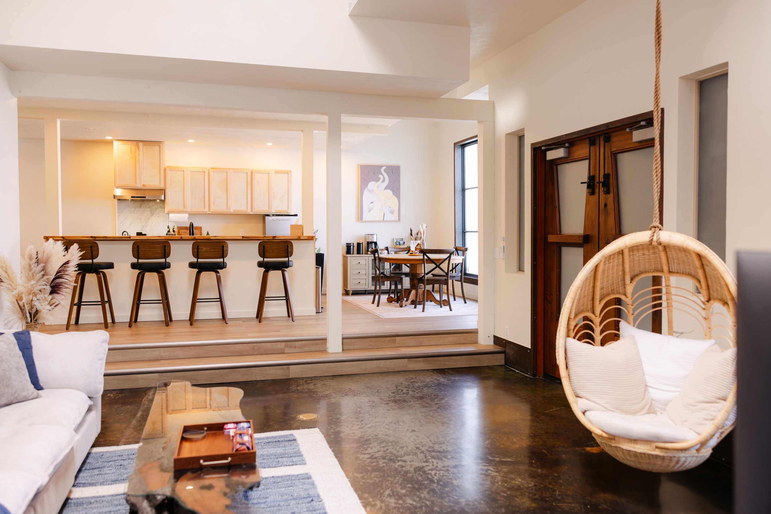Open-concept living and dining area with a kitchen island, bar stools, wooden dining table, and wicker chair, lit by natural light from large windows.