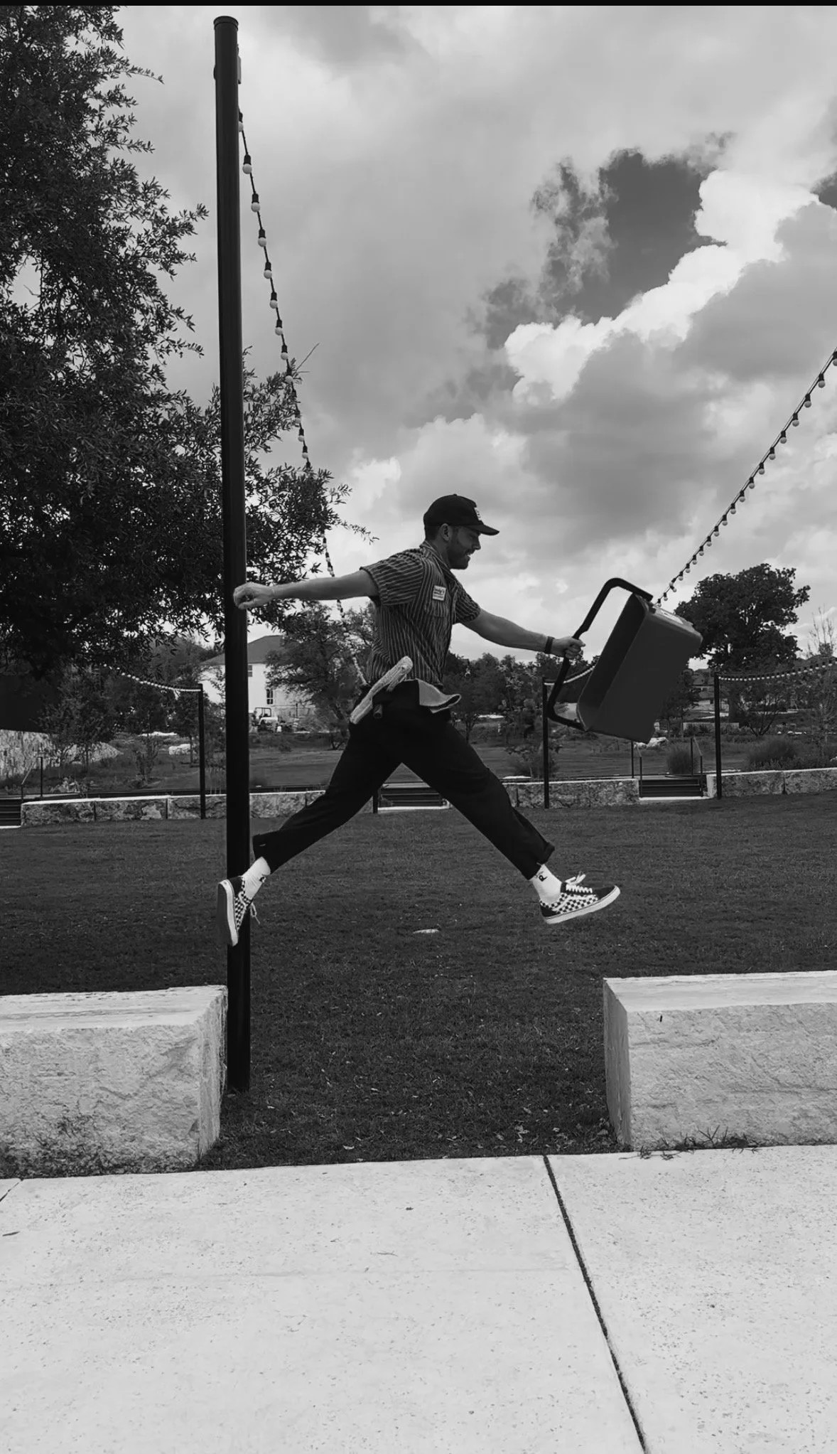 A man in a striped shirt, dark pants, striped Vans shoes, and a cap is jumping in the air outdoors, holding a chair and a roll of paper, with a cloudy sky, trees, and a park in the background.