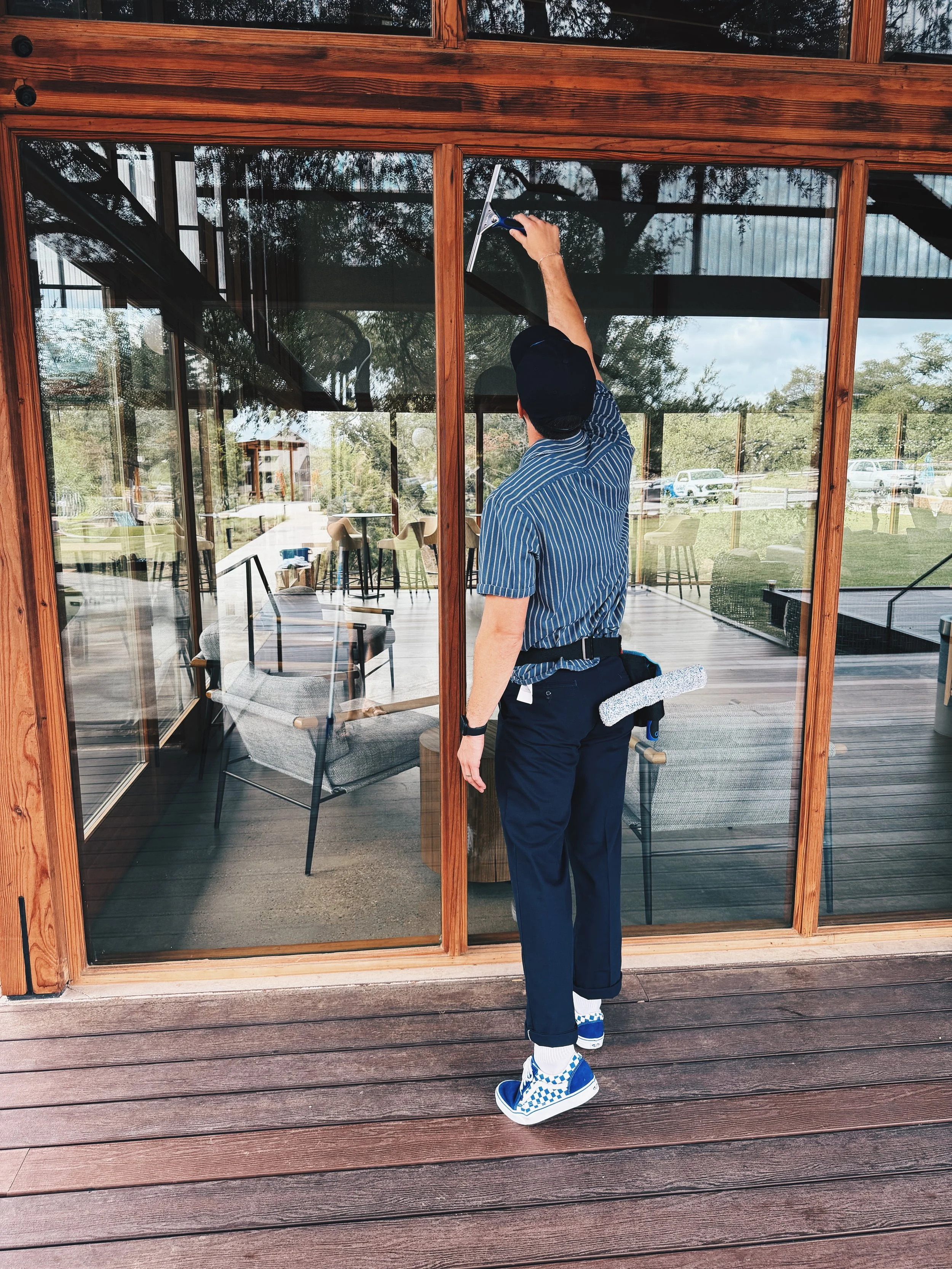 A person cleaning glass windows of a wooden-framed building with a squeegee, standing on a wooden deck, wearing sneakers and dark pants.