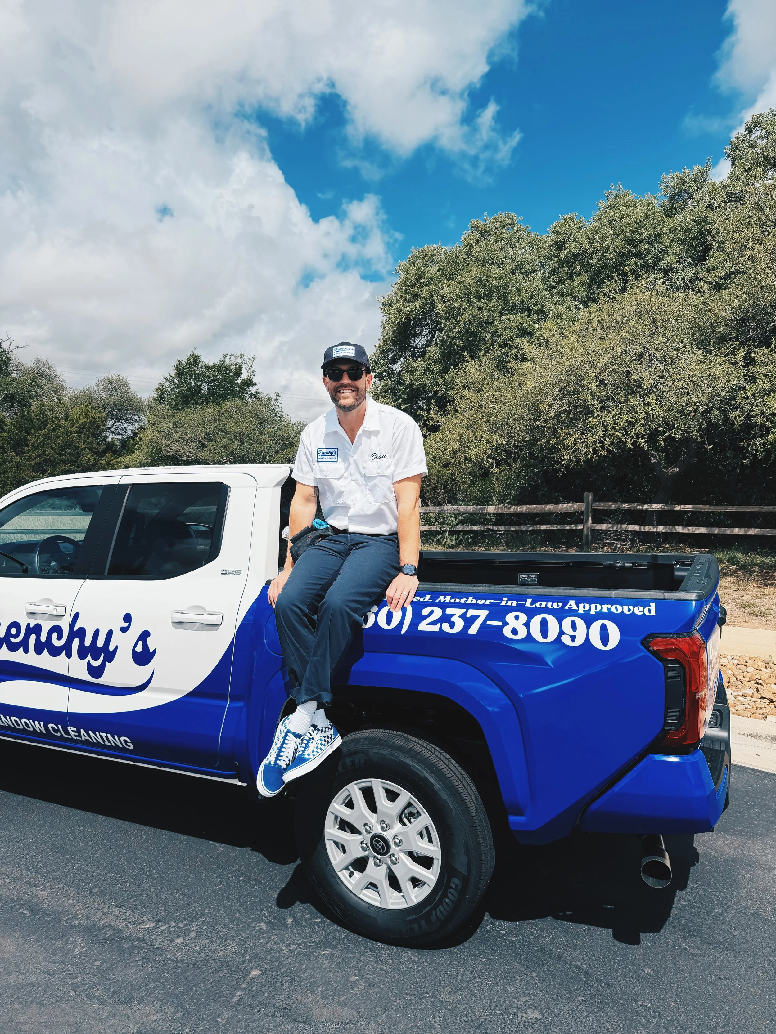 A man sitting on the side of a blue and white window cleaning company's pickup truck, smiling at the camera, outdoors with trees and a partly cloudy sky in the background.