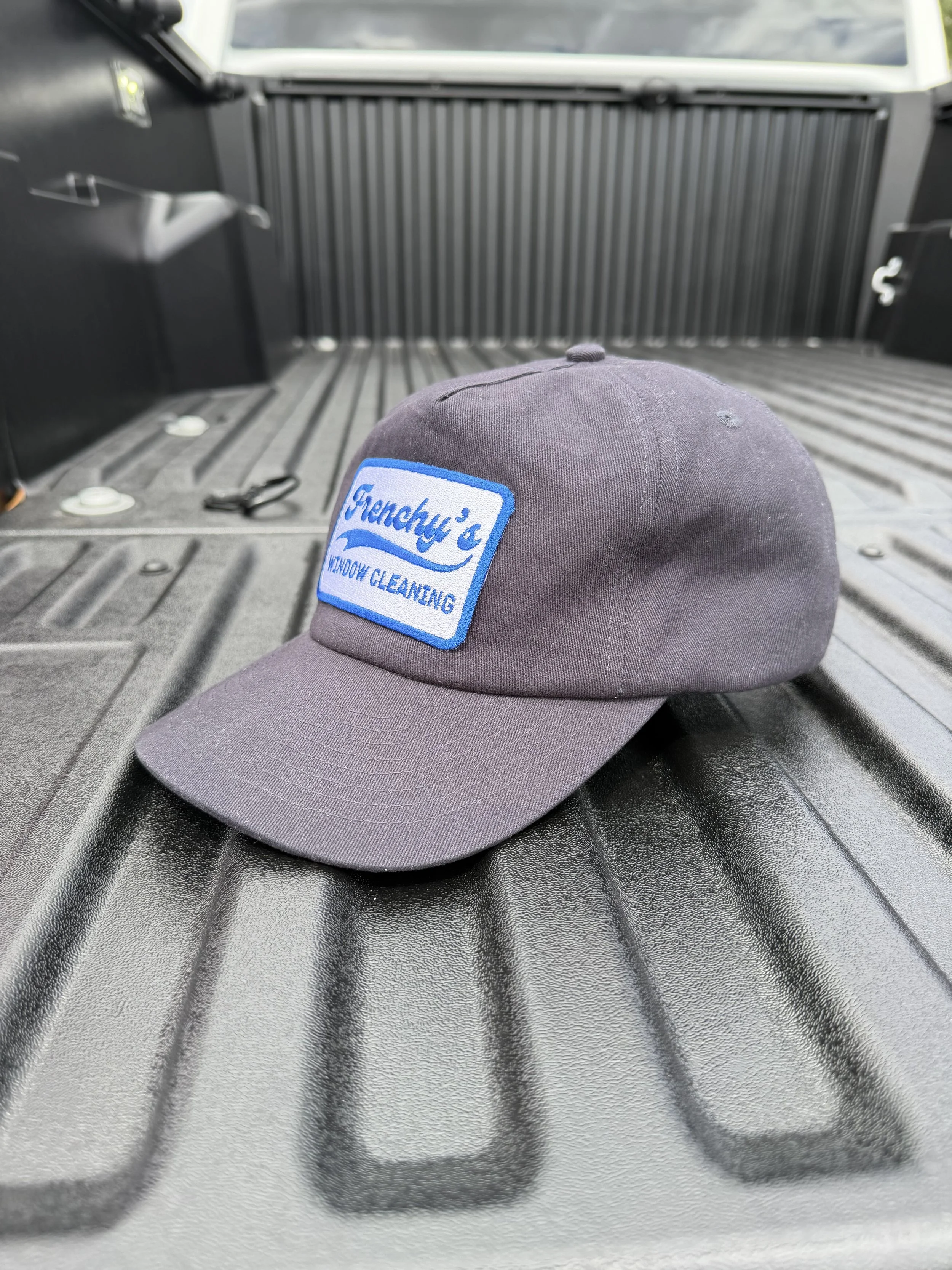 Gray cap with a blue and white patch that reads "Gretchy's Window Cleaning" placed on a truck bed with tools nearby.