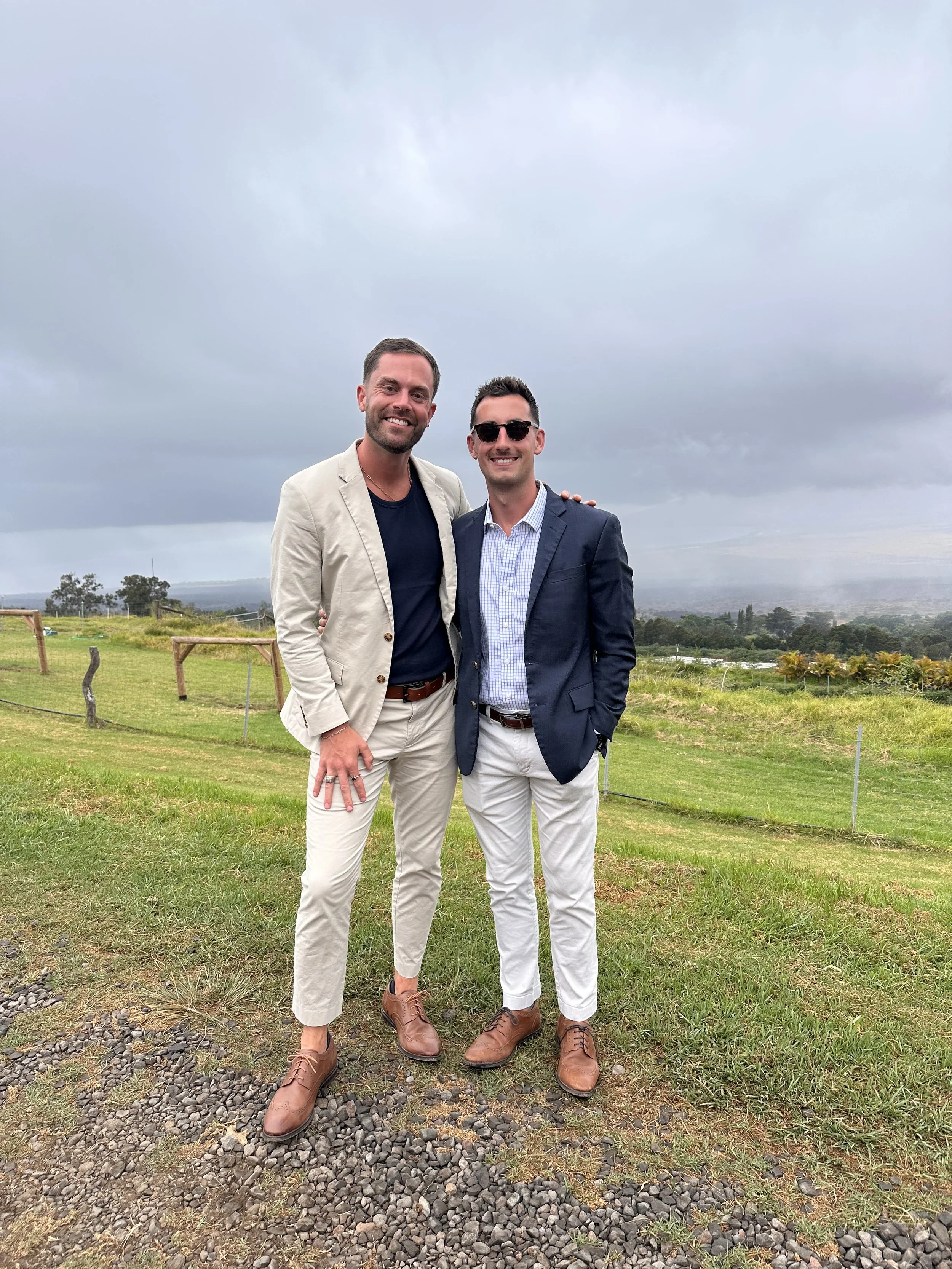 Two men dressed in suits standing outdoors on a grassy field, smiling at the camera, with a cloudy sky and rolling hills in the background.