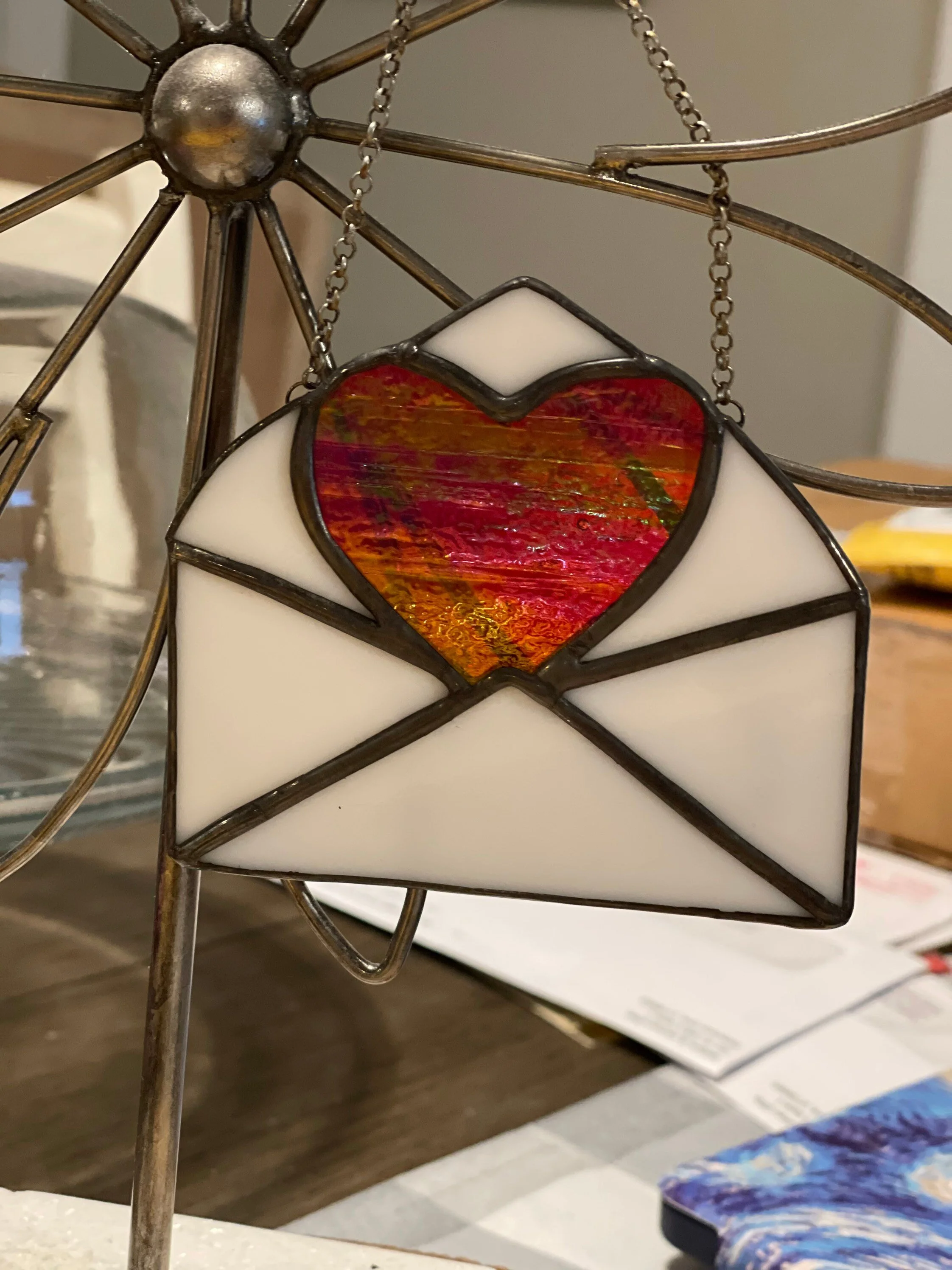 A stained glass lantern with a white exterior and a red heart design in the center, hanging from gold-colored chains attached to a metal frame.