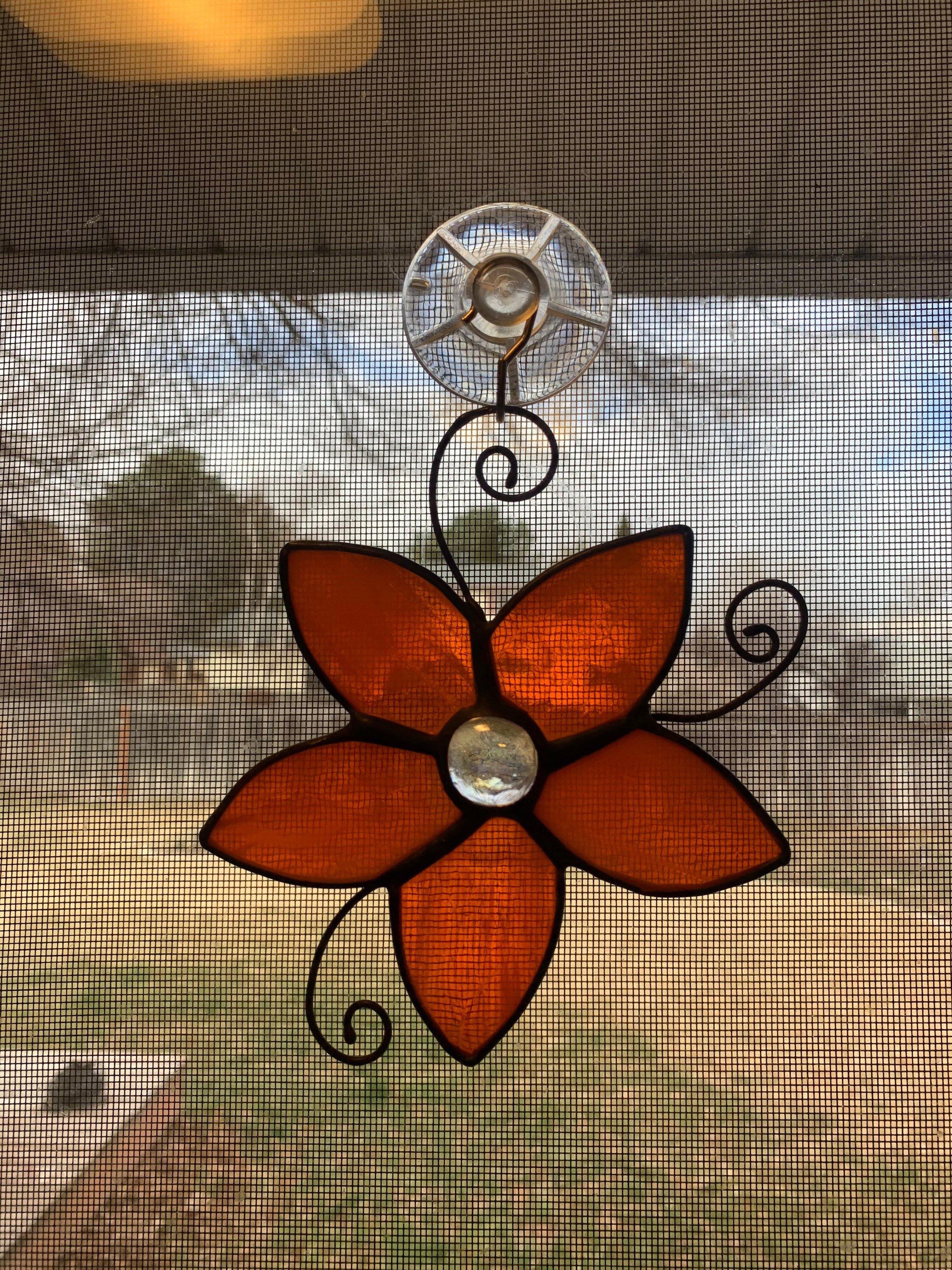 Close-up view of a stained glass butterfly decoration hanging in front of a window mesh screen, with a blurred outdoor background.