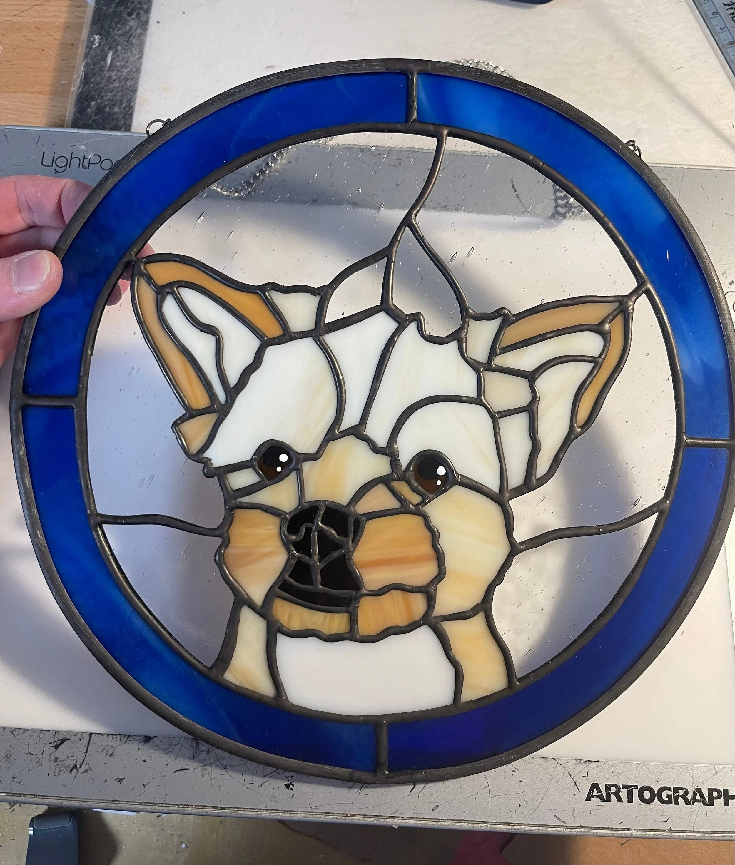 A stained glass art piece depicting a stylized dog face with a blue circular border, held over a work surface with a hand visible on the left side. The work surface has the word "ARTOGRAPH" printed on it.