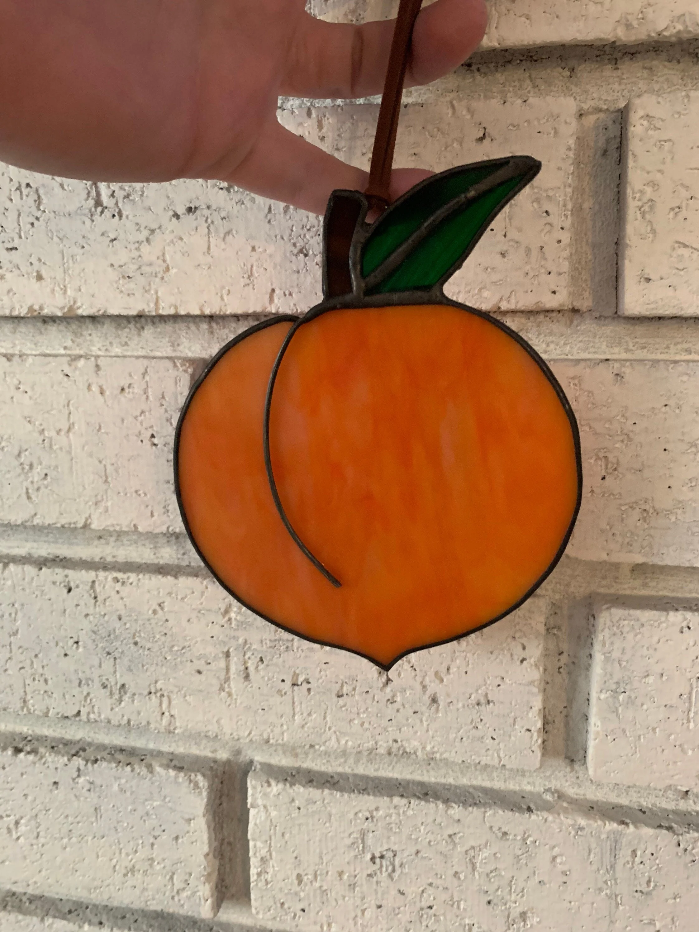 A stained glass art piece depicting an orange with a green leaf and stem, mounted on a white brick wall.
