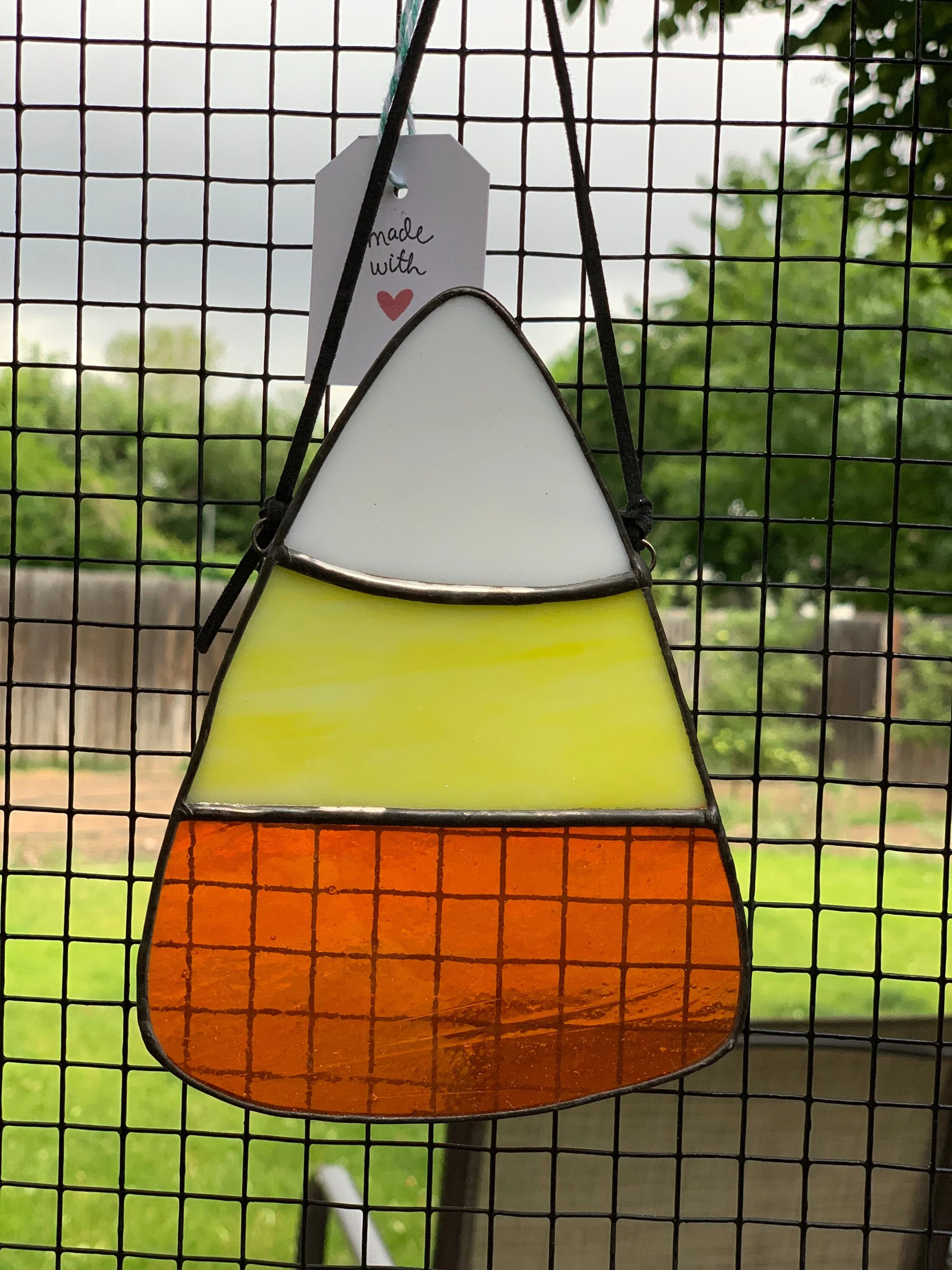 Colorful stained glass hanging ornament with a white top, yellow middle, and orange bottom, on a black wire frame grid, with a small card that says "made with" and a red heart, outdoors with trees in the background.