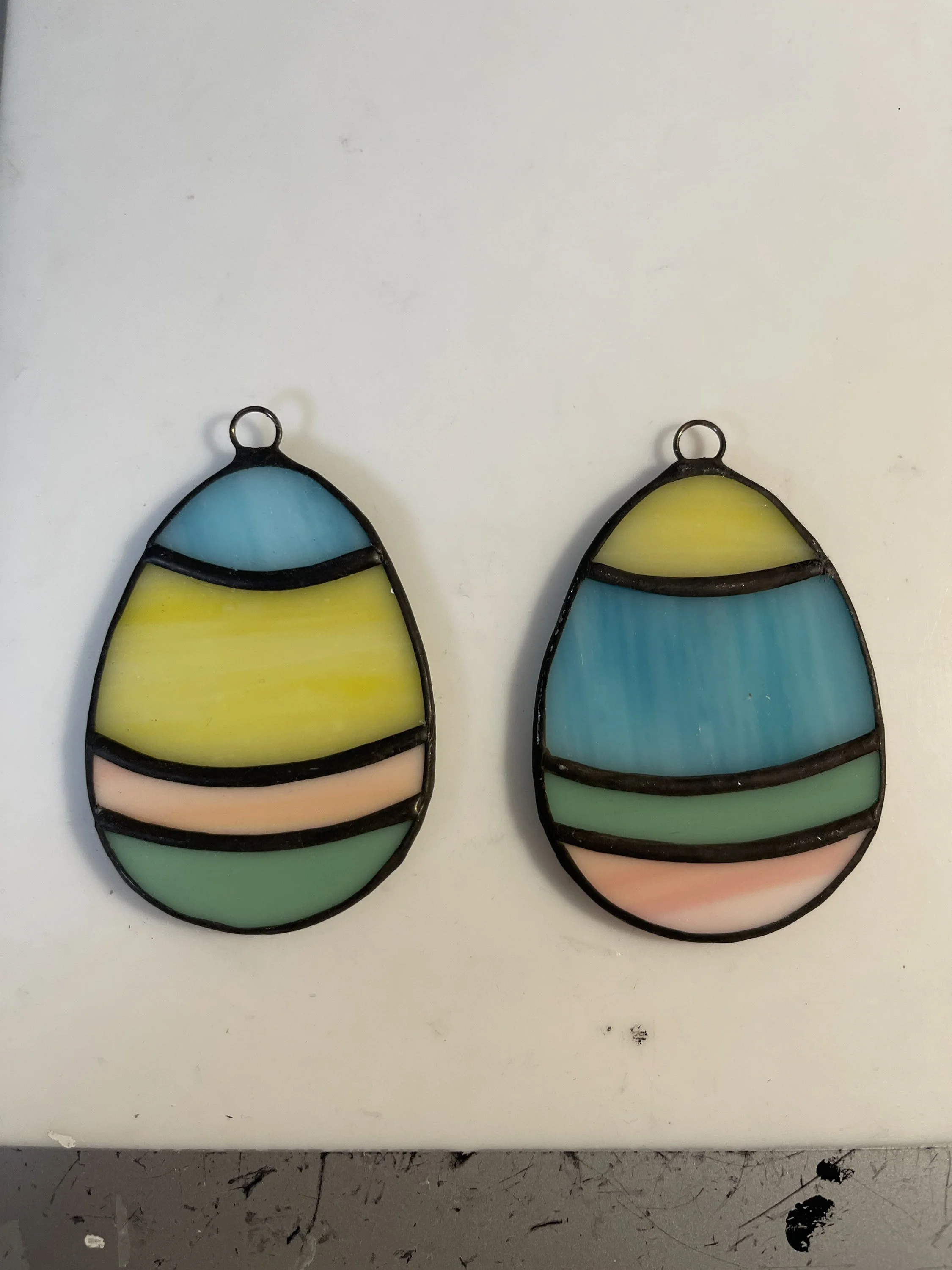 Two stained glass ornaments with colorful stripes, one with a blue, yellow, pink, and green pattern, and the other with a similar pattern in different shades, both hanging on a white surface.