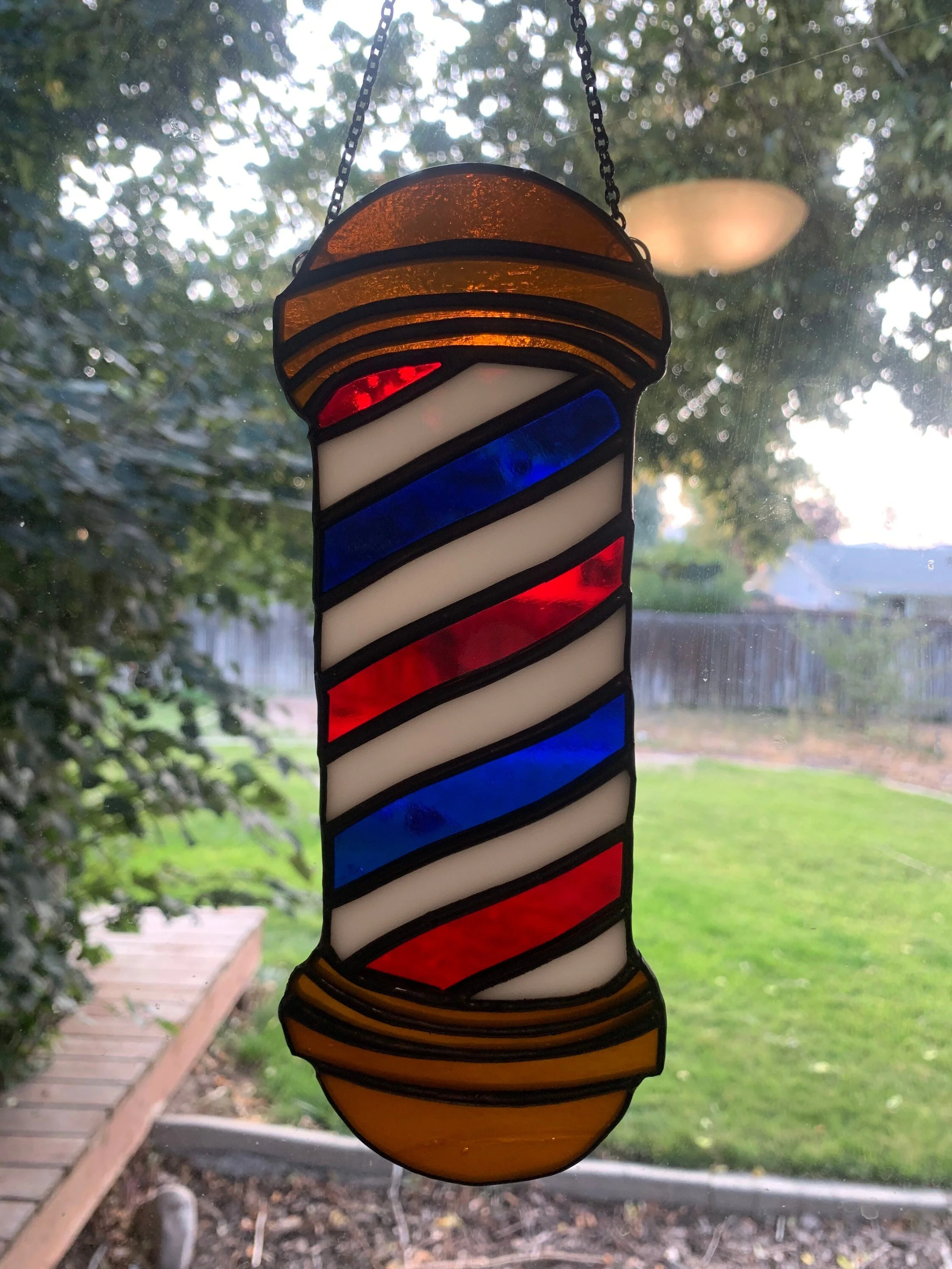 Colorful stained glass barber pole hanging in front of a window with a garden and trees outside.