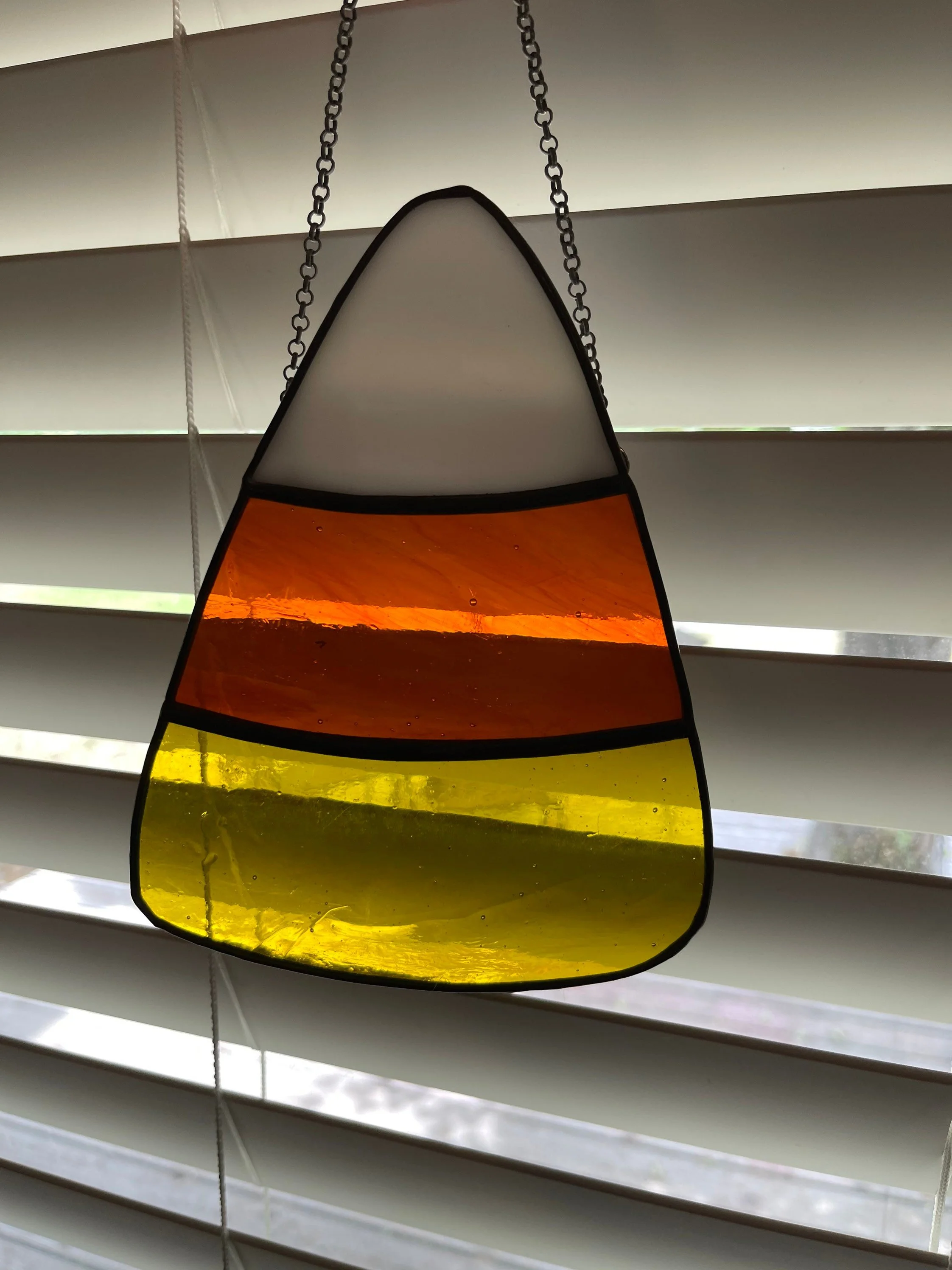 A stained glass hanging ornament with sections in yellow, orange, red, and white, suspended by chains in front of white window blinds.