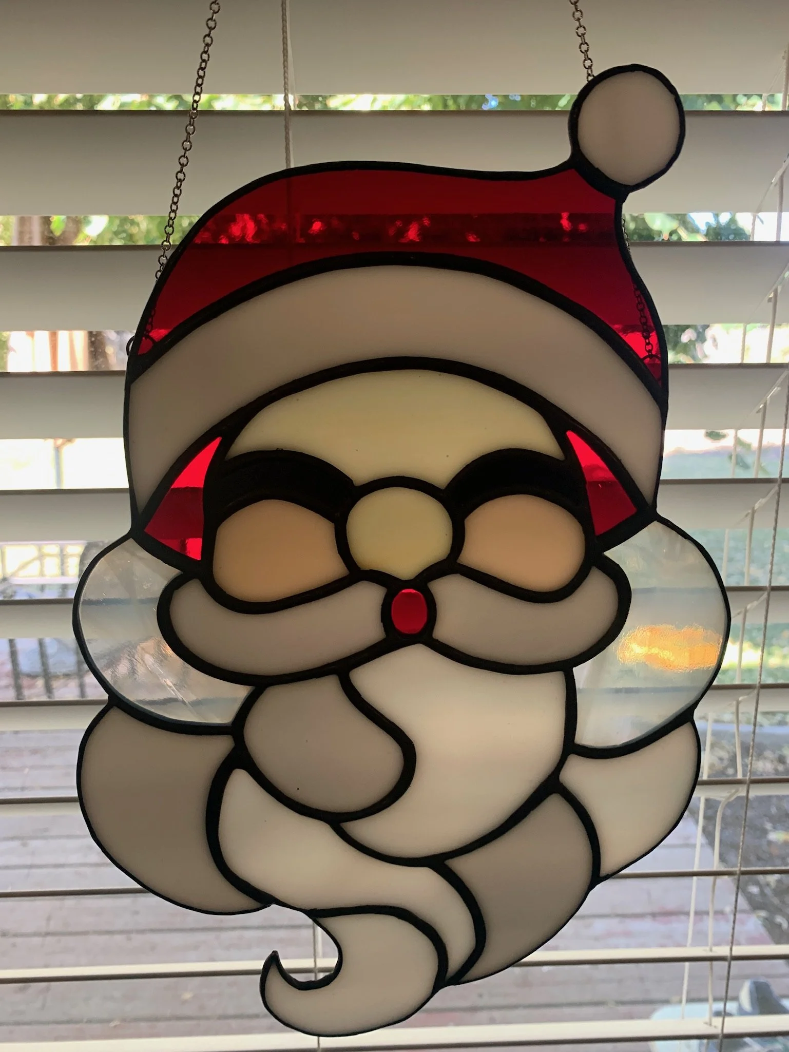 Decorative stained glass Santa Claus face hanging in front of window blinds with trees and blue sky outside.