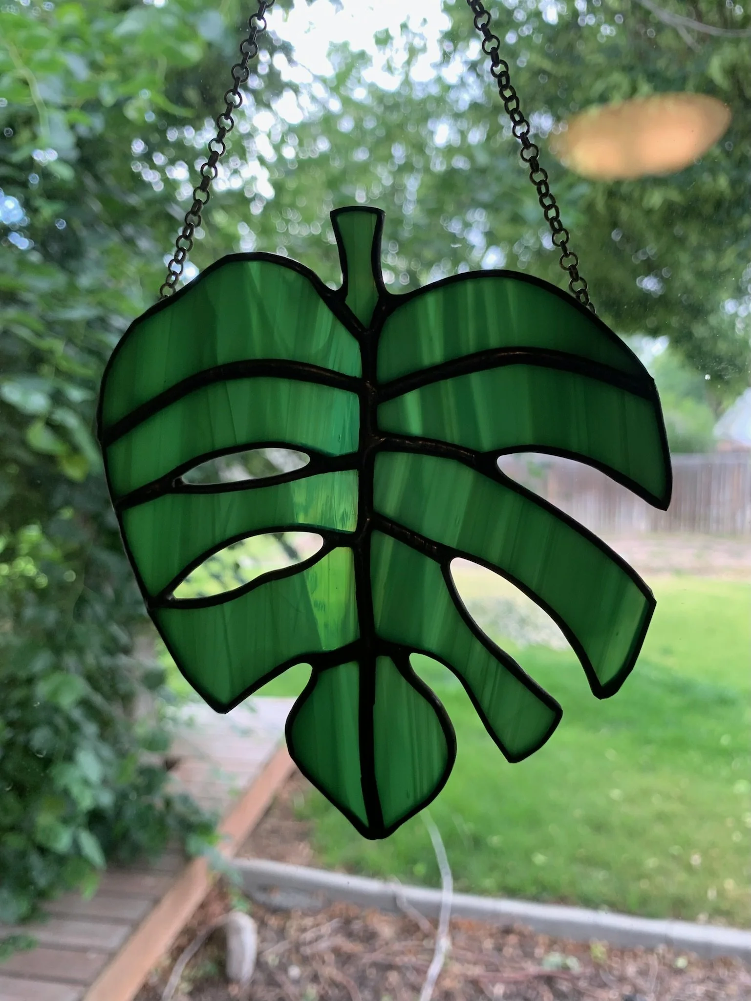 A green stained glass leaf-shaped wind chime hanging outdoors, with blurred greenery and a wooden walkway in the background.