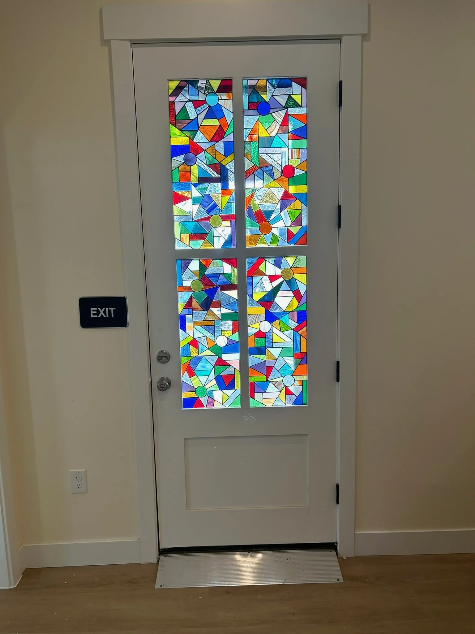 Colorful stained glass door with abstract geometric patterns and circles, and an EXIT sign on the wall to the left.
