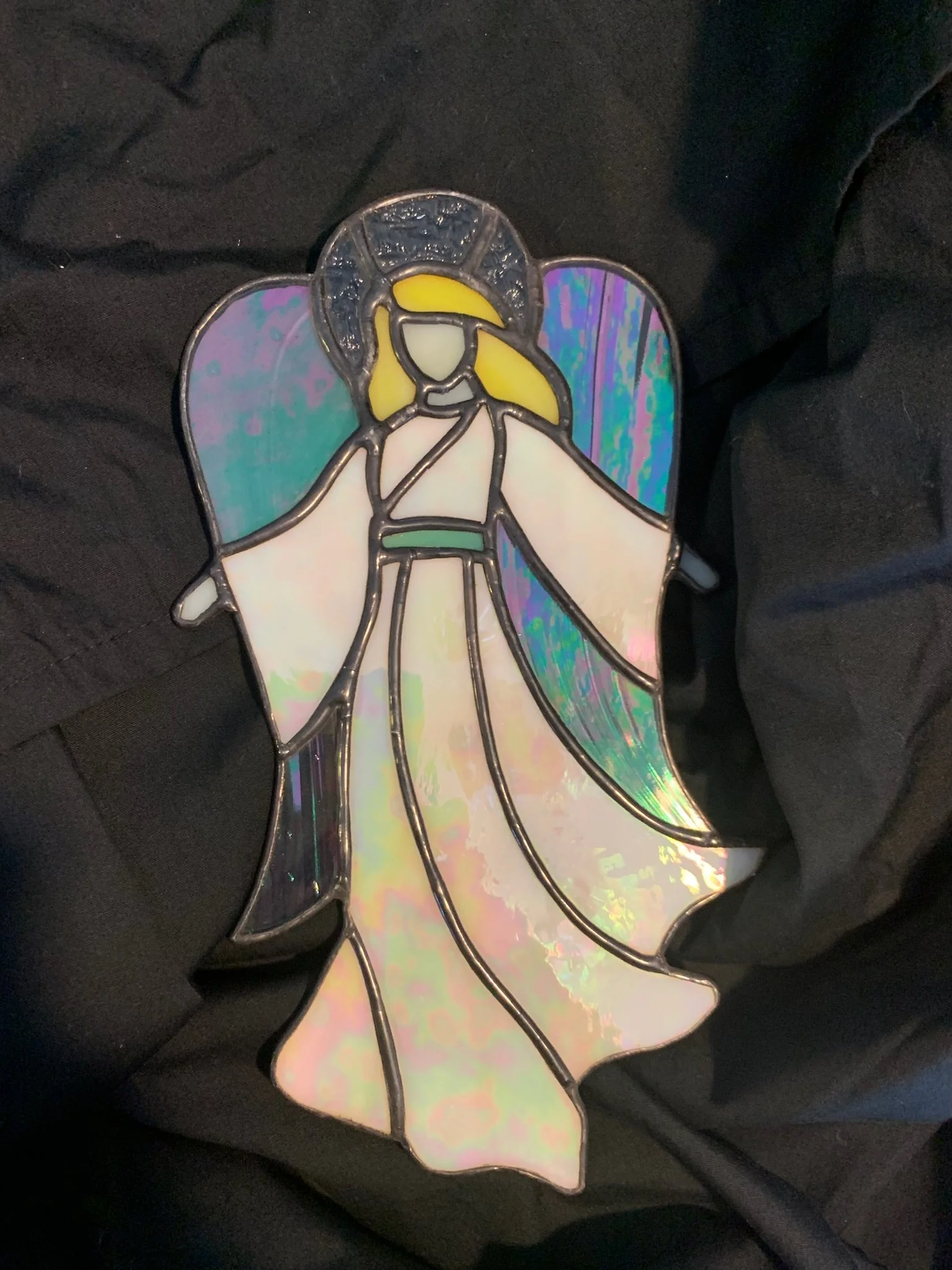 A stained glass angel pendant with iridescent wings and a halo, resting against a dark fabric background.