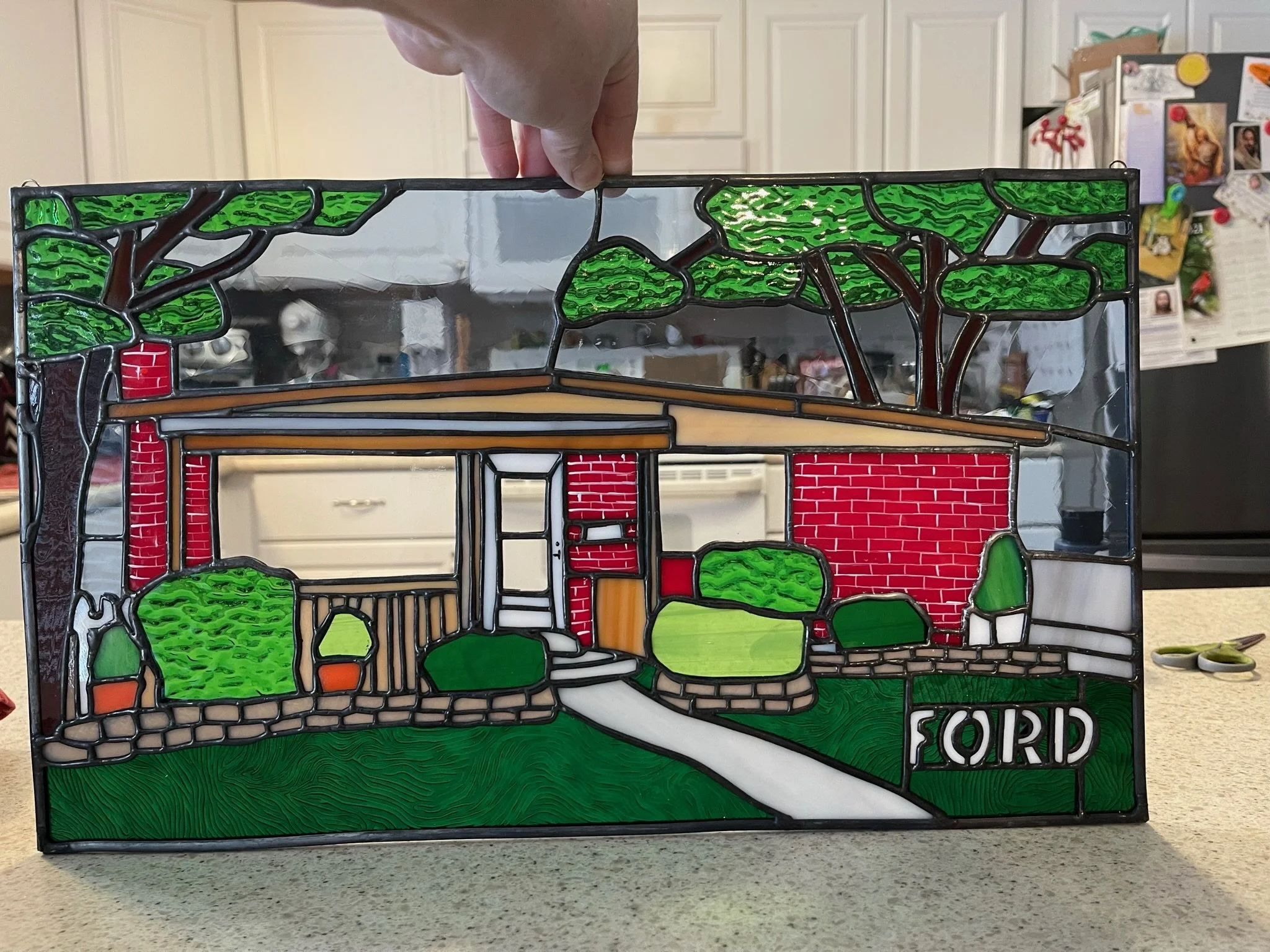 A colorful stained glass decorative panel depicting a house with trees, grass, and the text 'GRAND FORD' at the bottom.