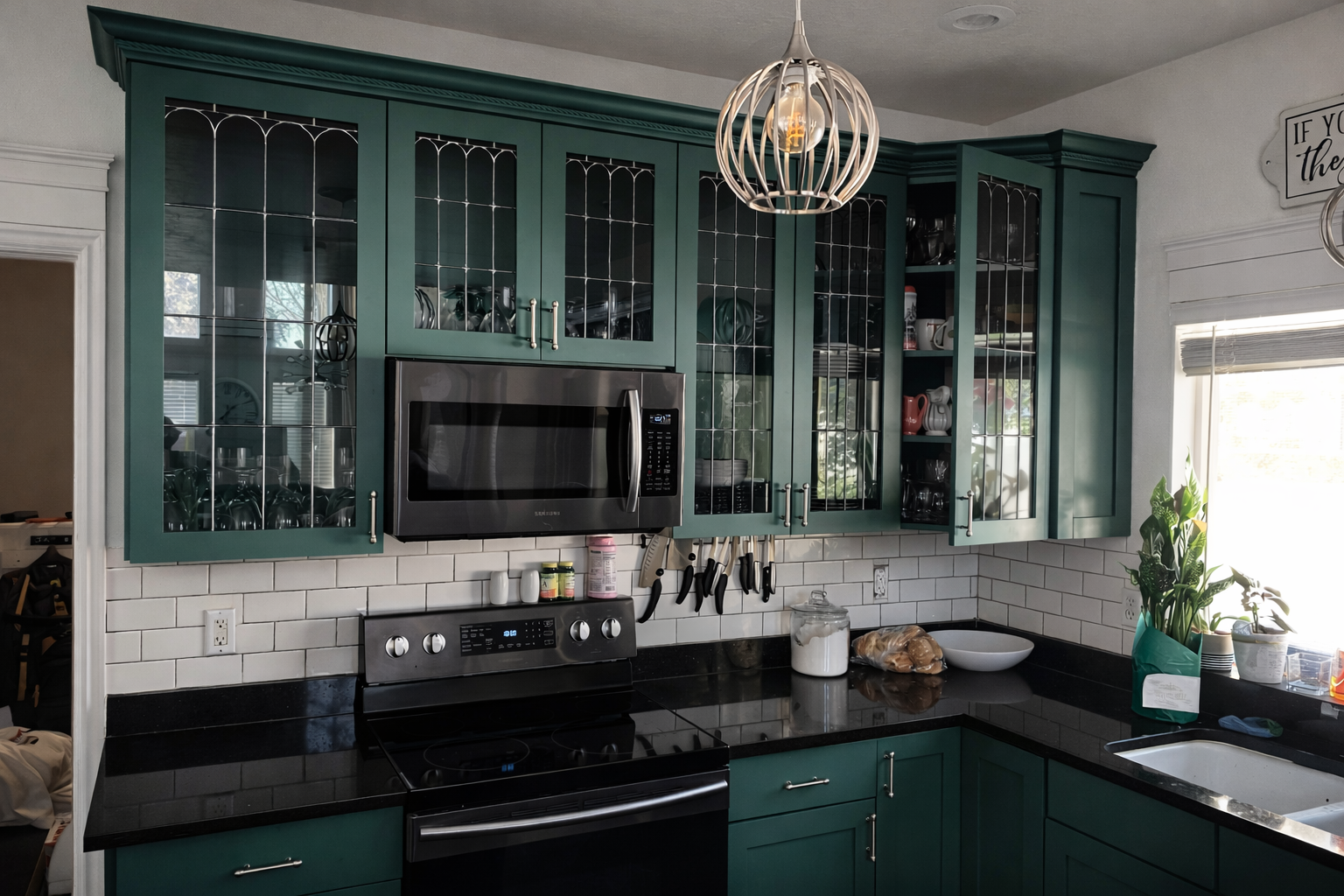 Kitchen with green cabinets, black countertop, white tiled backsplash, microwave, stove, and a window with a plant on the windowsill.