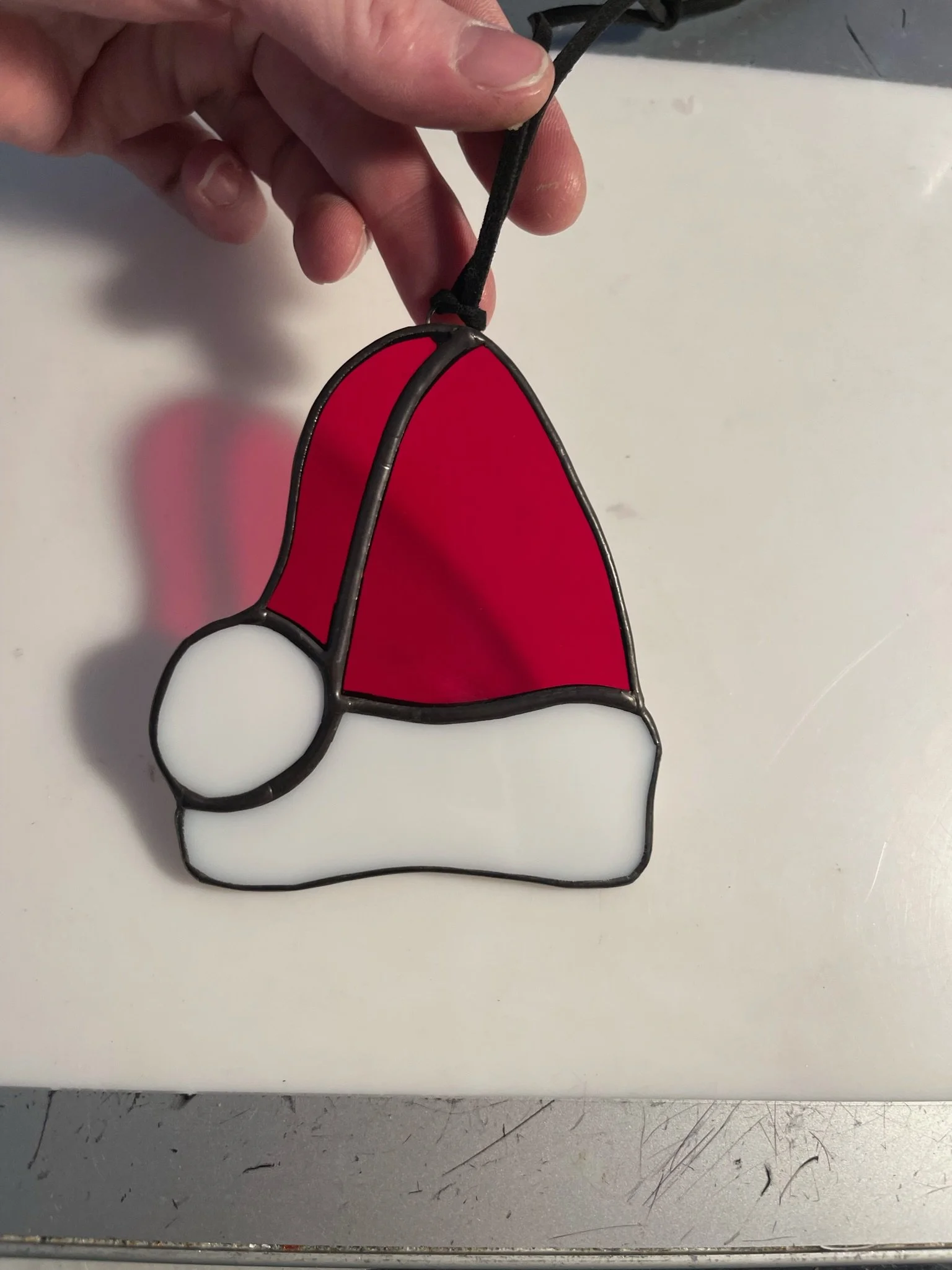 A stained glass ornament of Santa Claus wearing a red hat and suit with a white beard, held by a hand with a black string attached.
