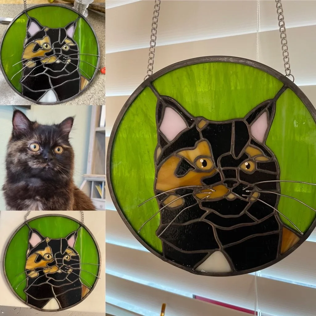 A stained glass pendant featuring a stylized cat face with yellow, black, and white glass, set against a green background, hanging from a chain.