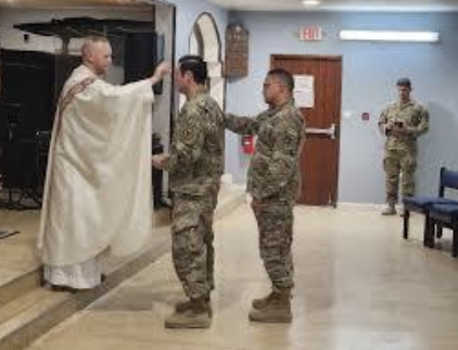 A priest performing a blessing for two soldiers in camouflage uniforms in a room, with a third soldier standing in the background.