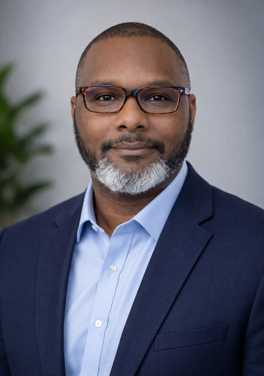 Close-up portrait of a confident African American man with a beard and glasses, wearing a navy suit and blue dress shirt, standing in front of a plain gray background.