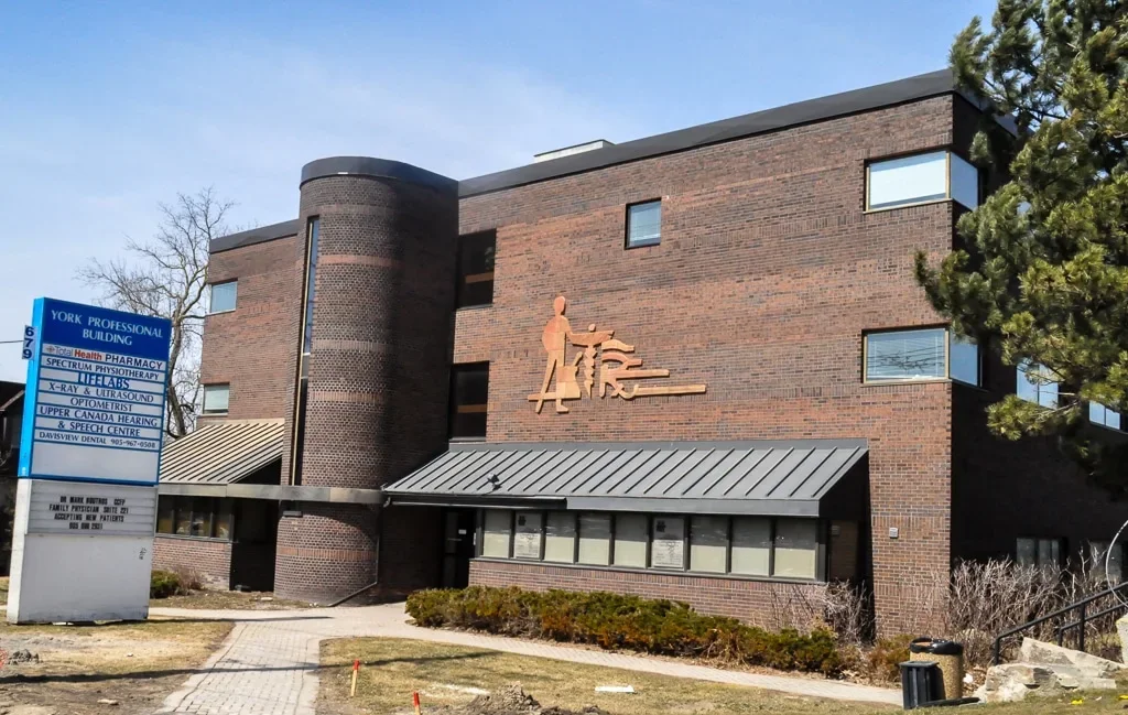 A multi-story brick building with a metal awning above the entrance and a large sign indicating it offers medical, dental, and audiology services. There is a small pathway leading to the entrance and a green shrubbery in front.
