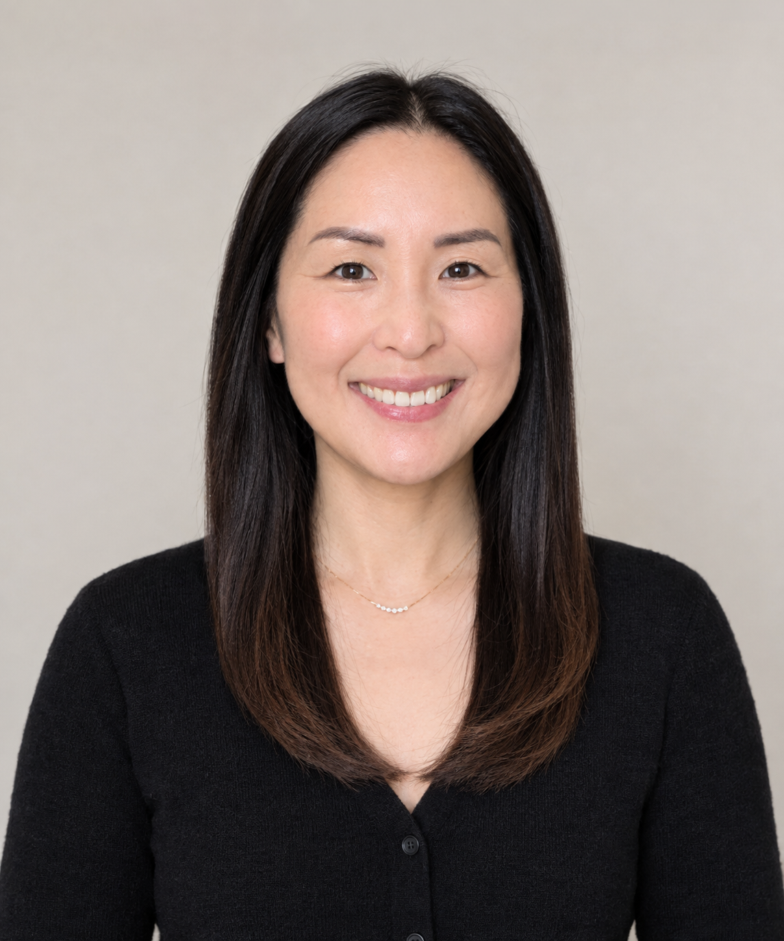 A woman with long black hair smiling, wearing a black top and a delicate necklace, against a plain background.