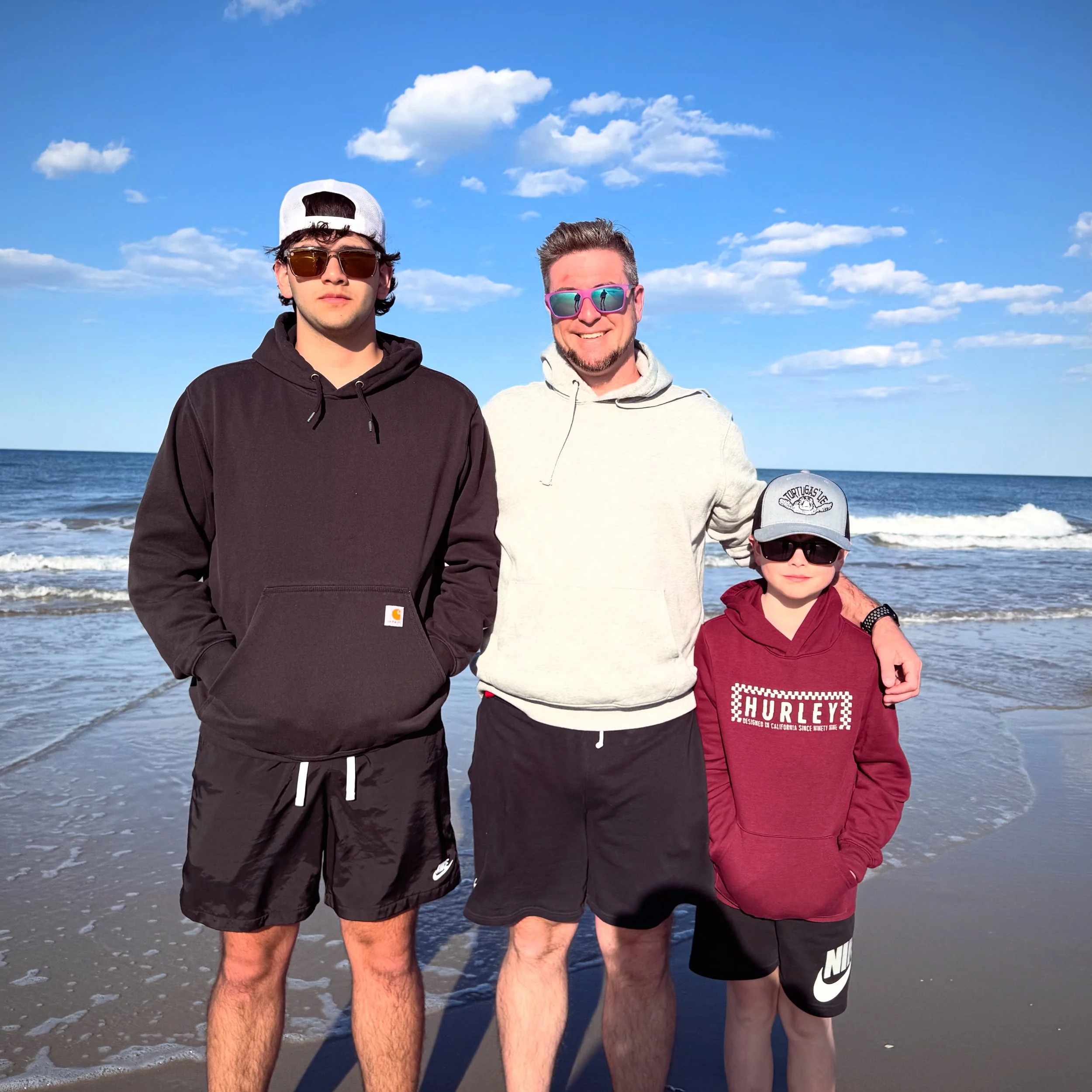 Matt with his 2 sons, standing on the beach near the ocean, wearing sunglasses and casual hoodies and shorts, with blue sky and clouds in the background.