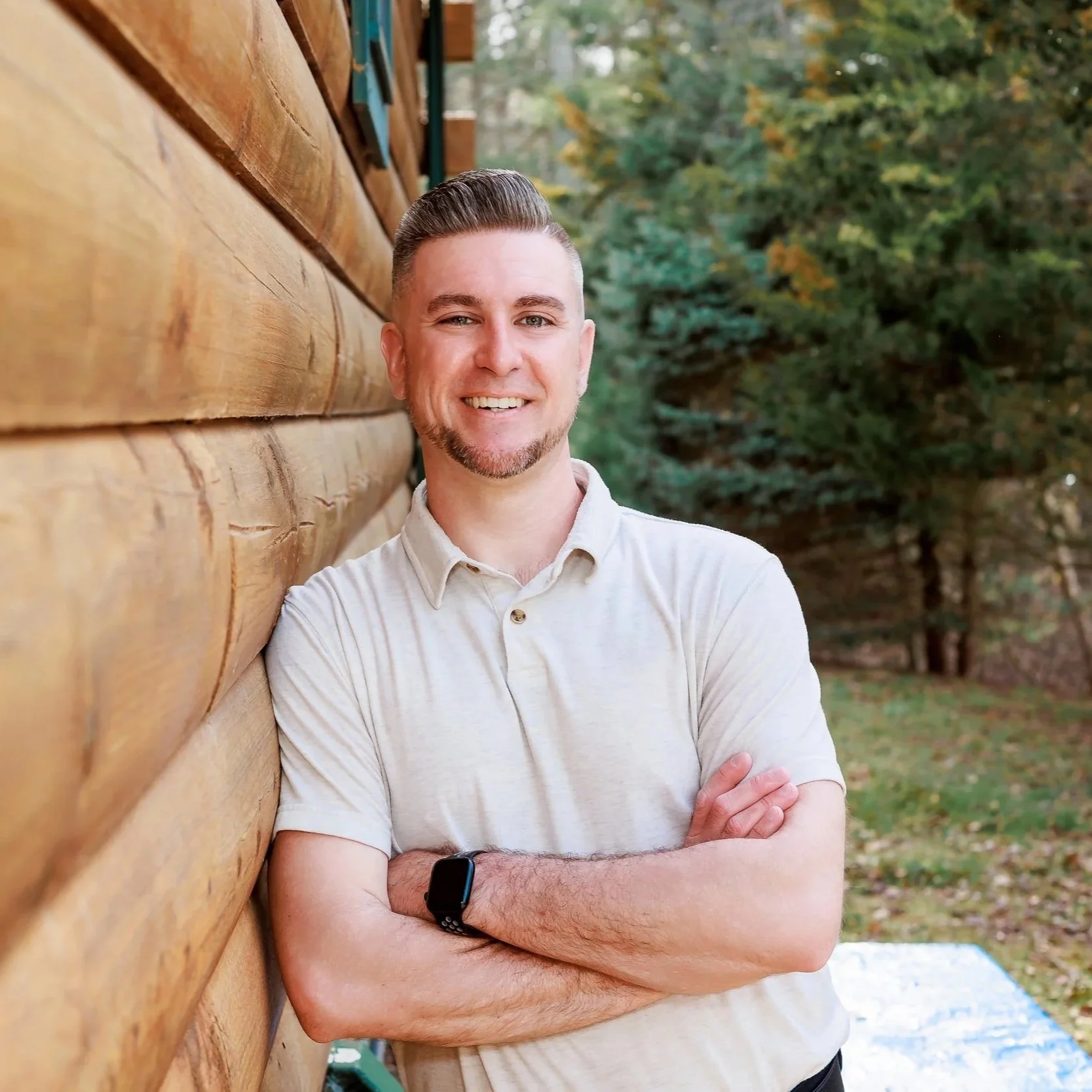 Matt, wearing a light-colored polo shirt and a smartwatch on his wrist, standing outdoors against a wooden wall with trees in the background.