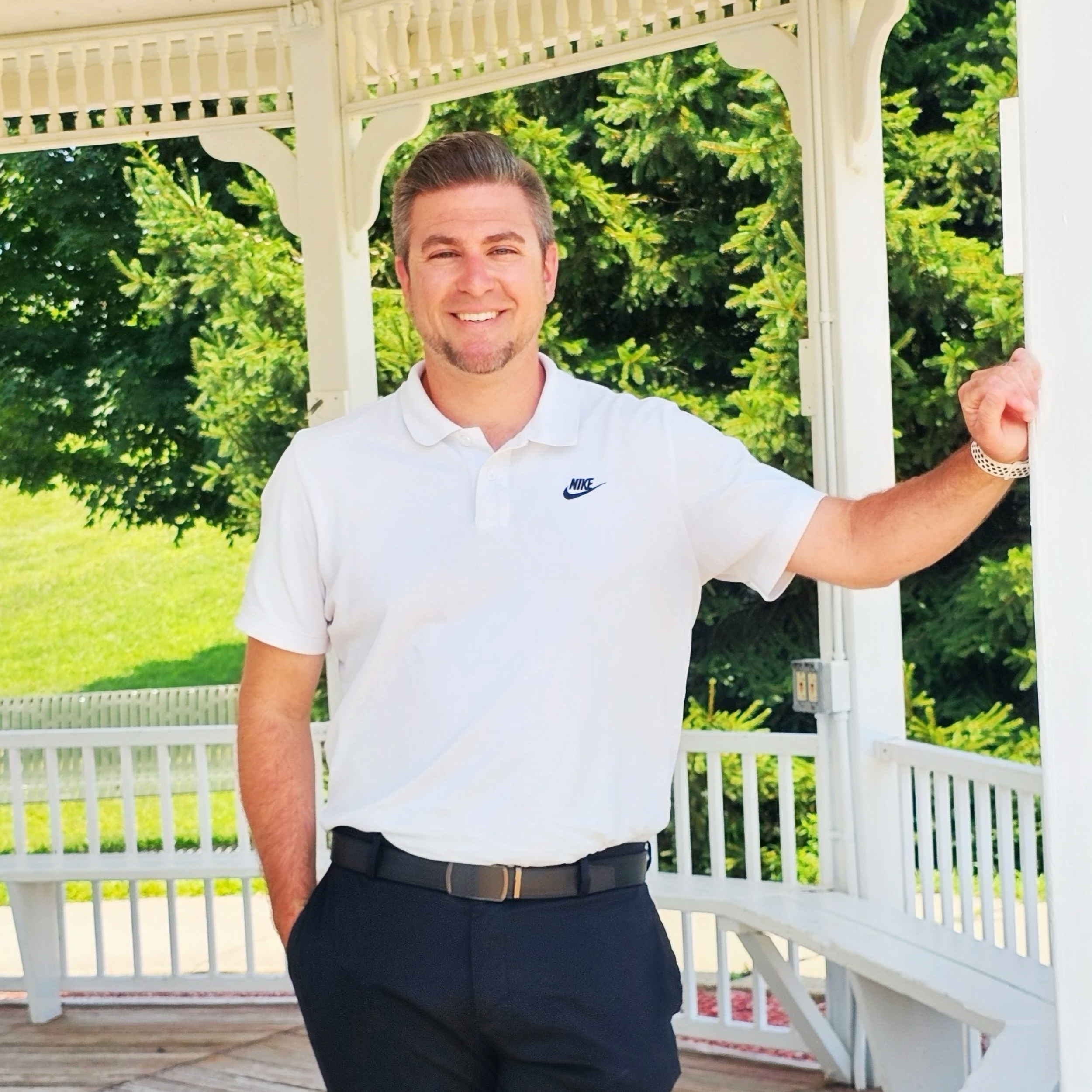 Matt in a white collared shirt standing outdoors in front of a white gazebo with green trees in the background, smiling.