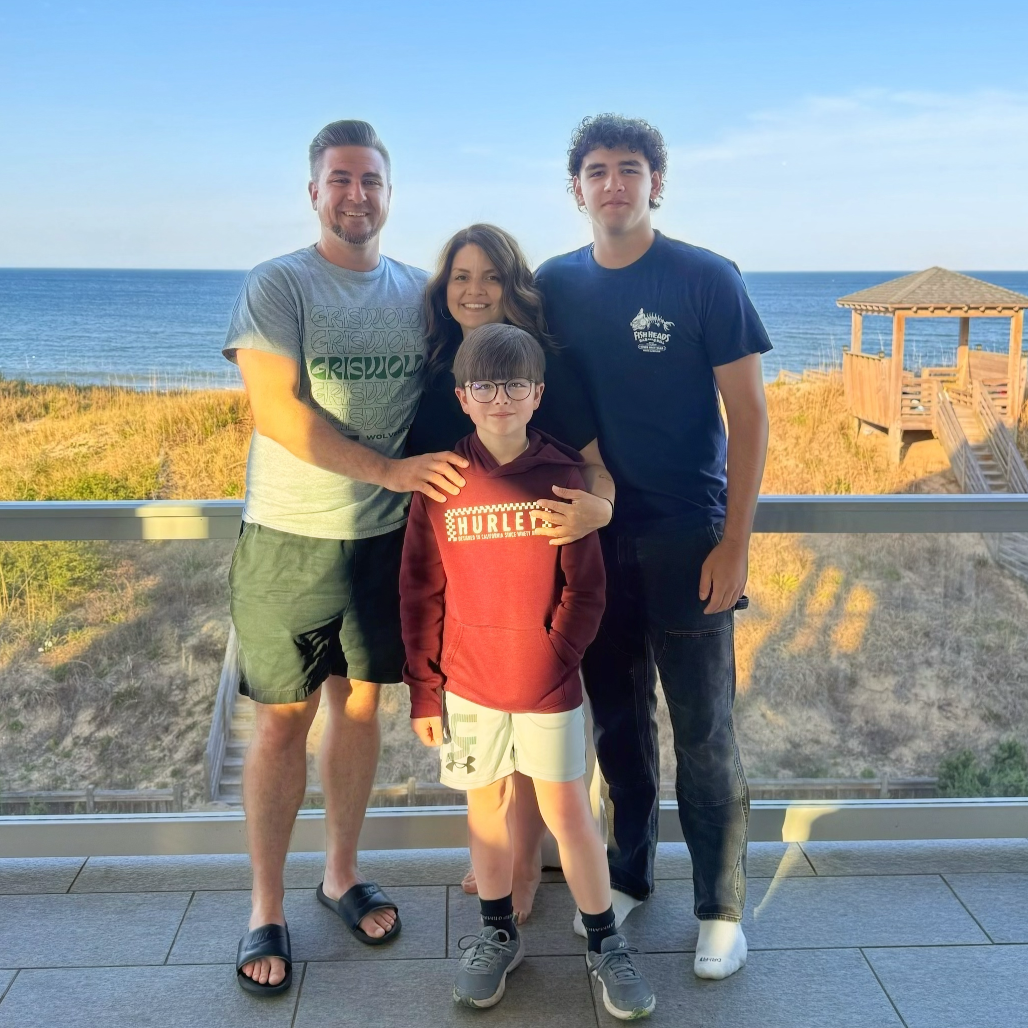 Matt is next his wife and 2 sons, standing outdoors by the ocean on a wooden deck with a small gazebo in the background, during daylight. They are smiling and posing for the photo.