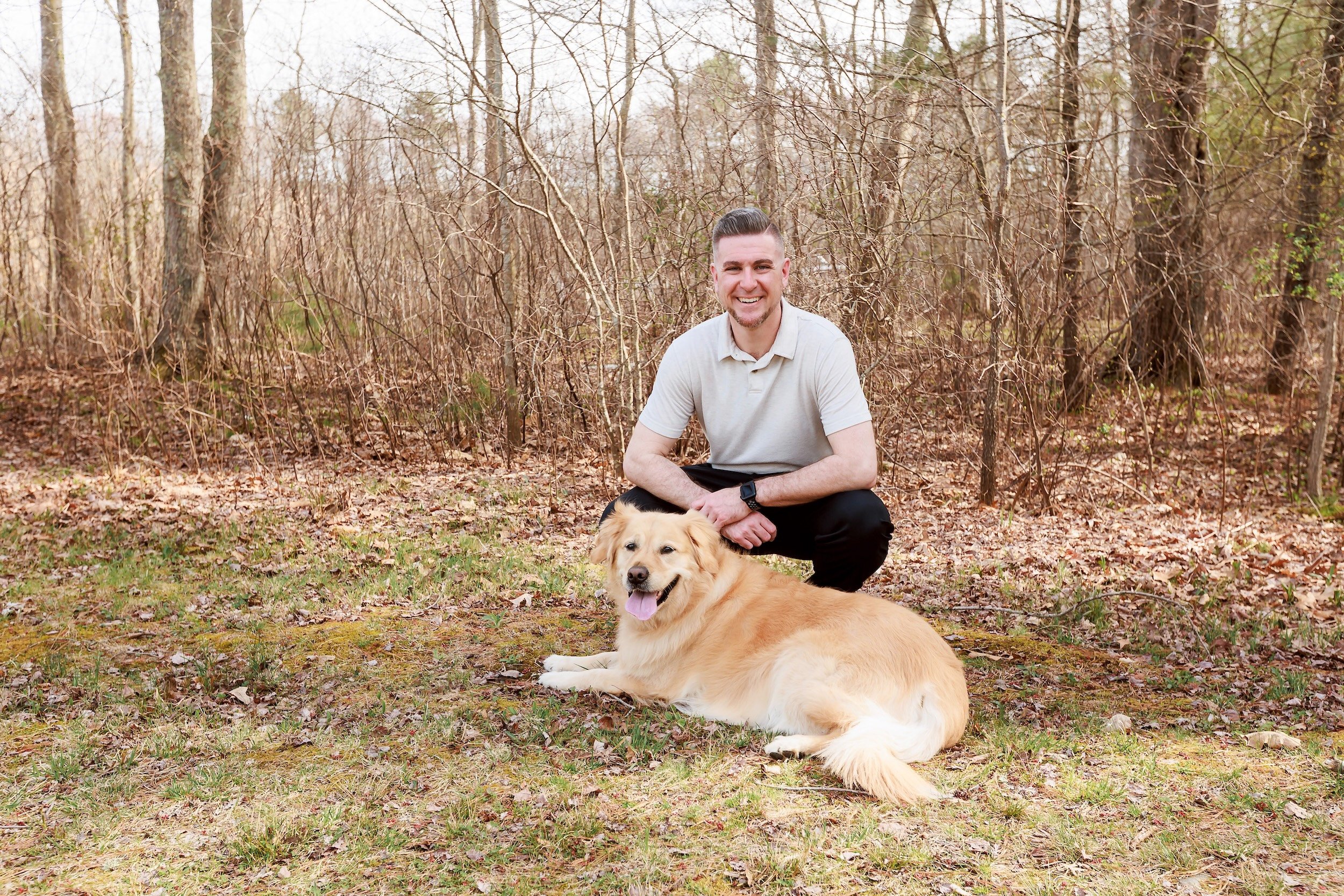 Matt kneeling next to a golden retriever lying on the ground in a wooded area with leafless trees.