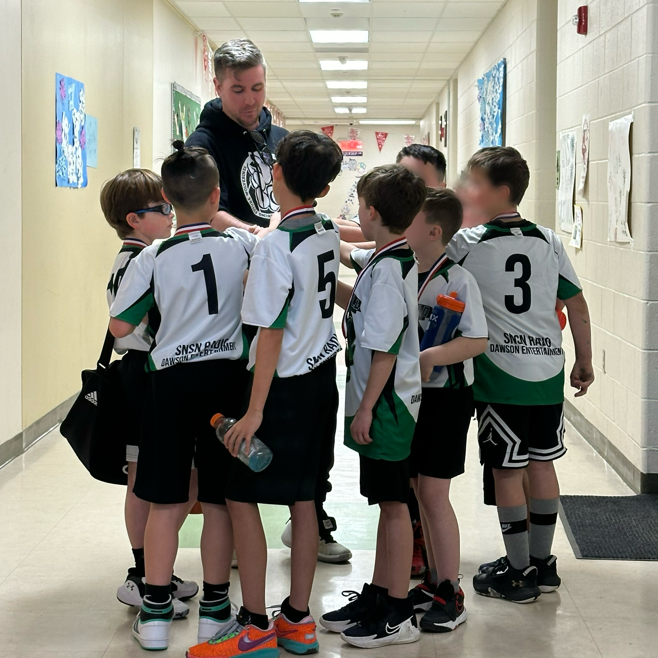 A group of young boy basketball players wearing jerseys, standing in a huddle with Matt, their coach in a hallway decorated with red banners and sports-themed artwork.