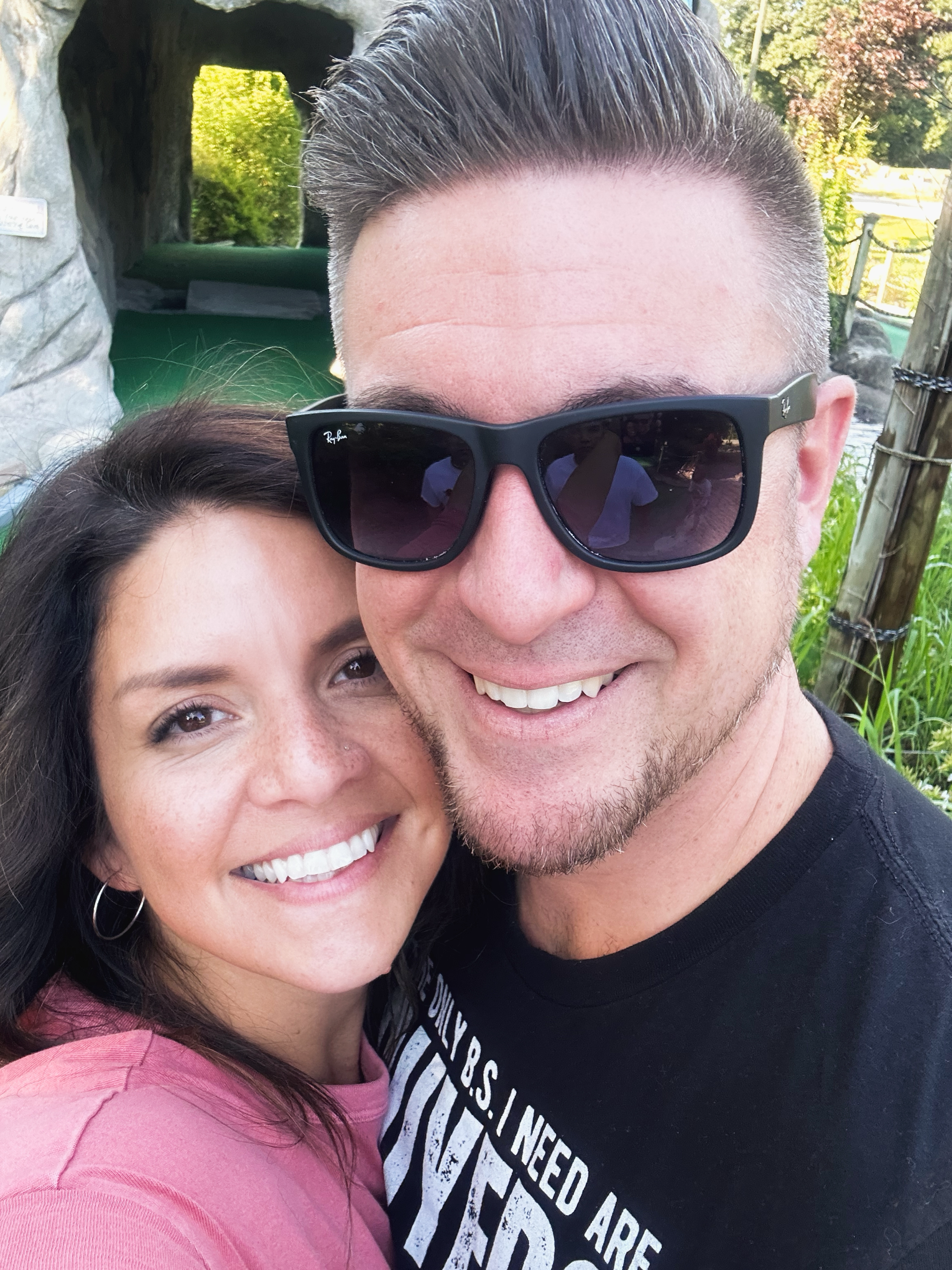 Close-up of Matt and his wife taking a selfie outdoors. The woman has long dark hair, and the man is wearing black sunglasses and has short, styled hair.