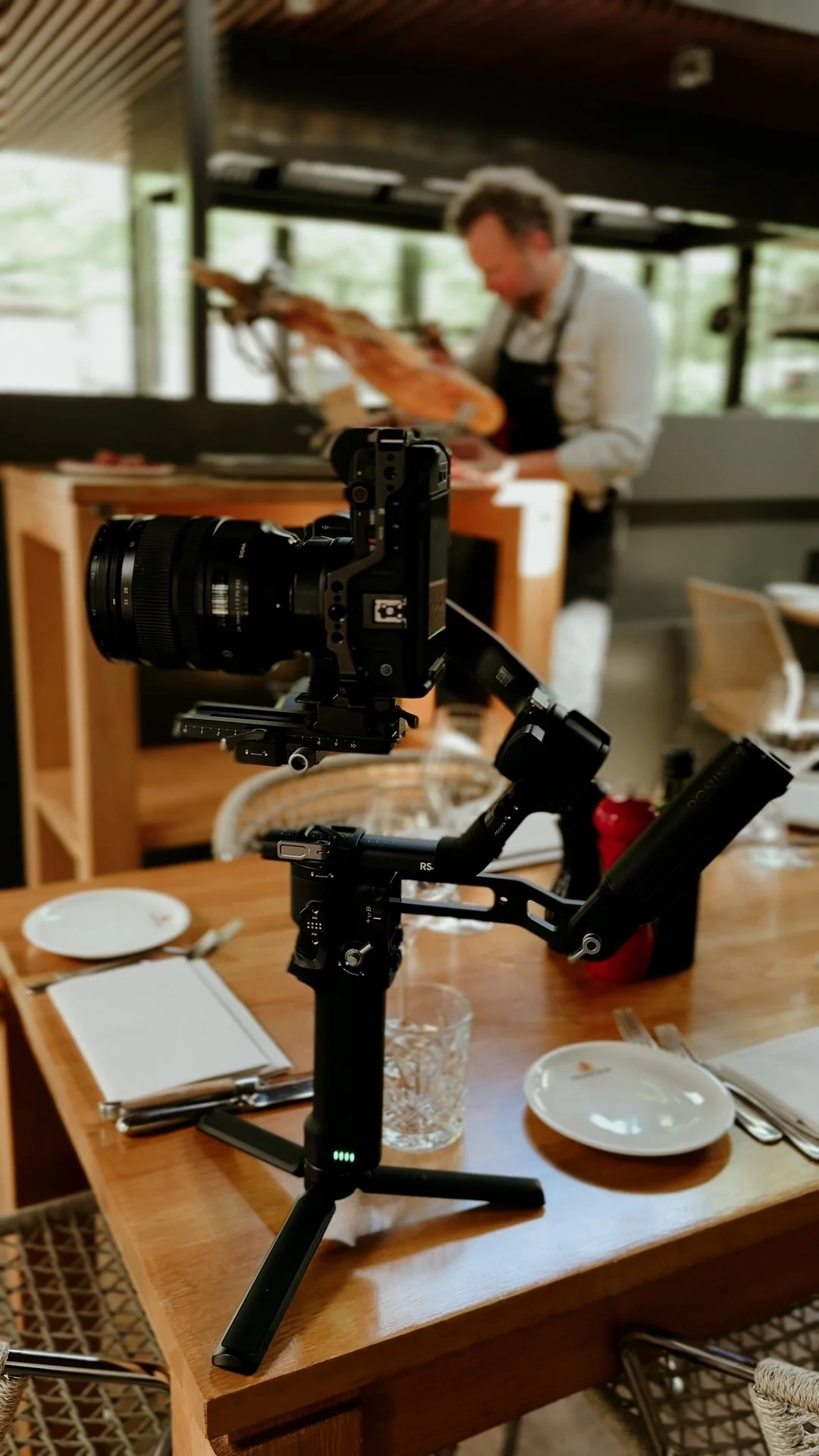 een camera op een stabiele standaard gericht op een tafel met eten en drinkgerei, en een chef-kok die op de achtergrond een gerecht bereidt in een keuken of restaurant.