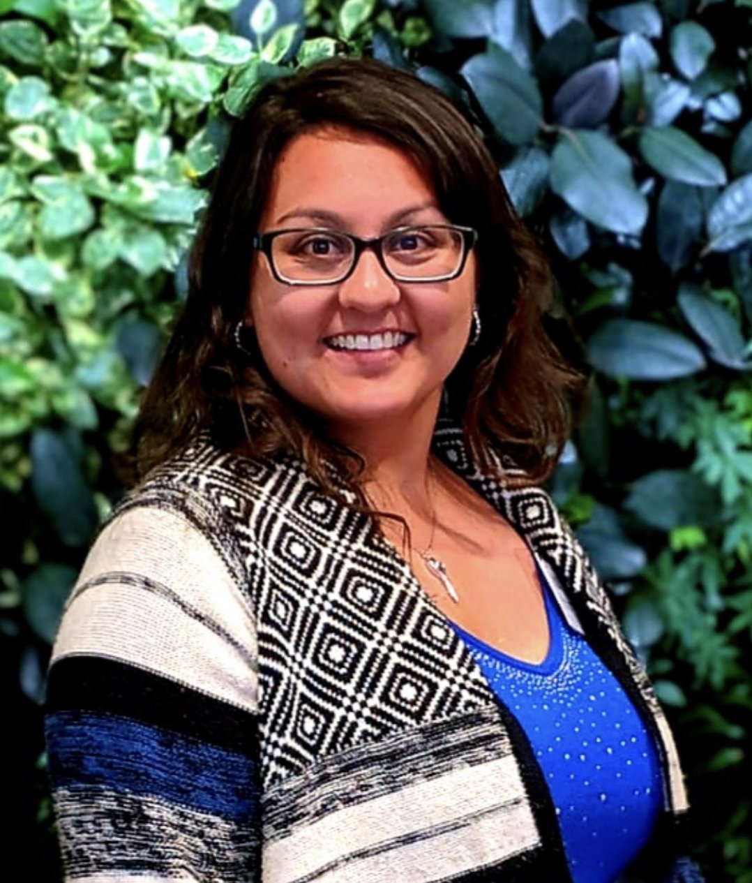 A smiling woman with glasses and dark brown hair, wearing a patterned jacket over a blue top, standing in front of green leafy plants.