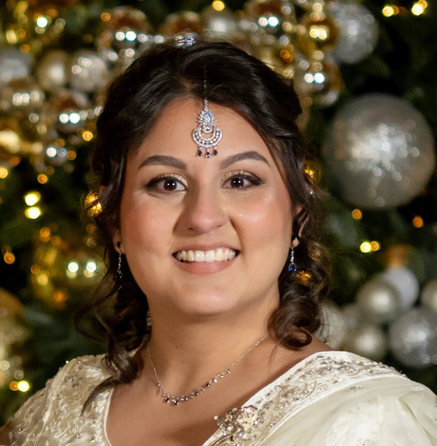 A woman with dark curly hair, wearing jewelry and a white dress, smiling in front of a decorated Christmas tree with gold and silver ornaments and lights.