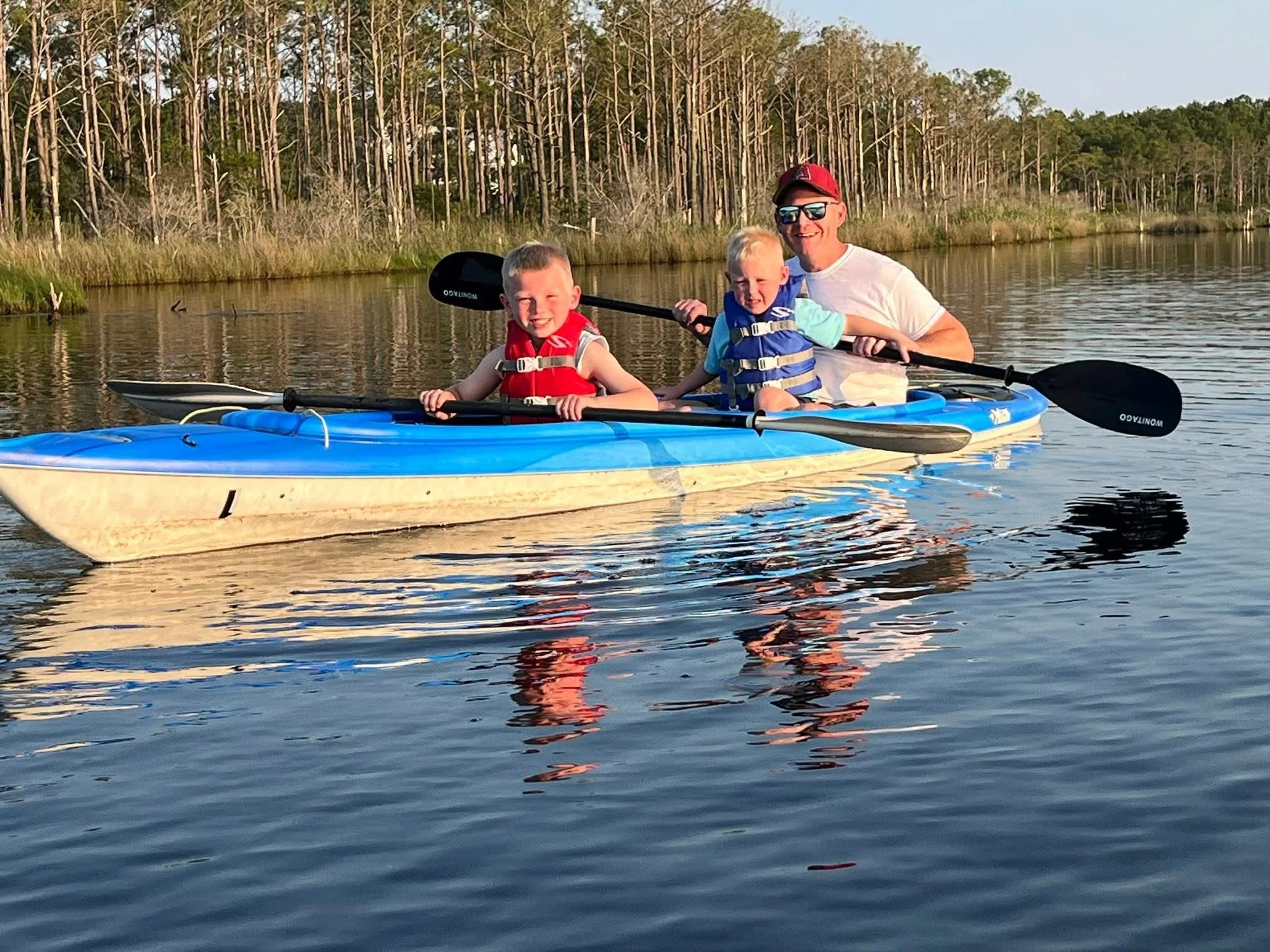 Two children and an adult are kayaking on a calm body of water, wearing life jackets, with a backdrop of tall trees and a clear sky.