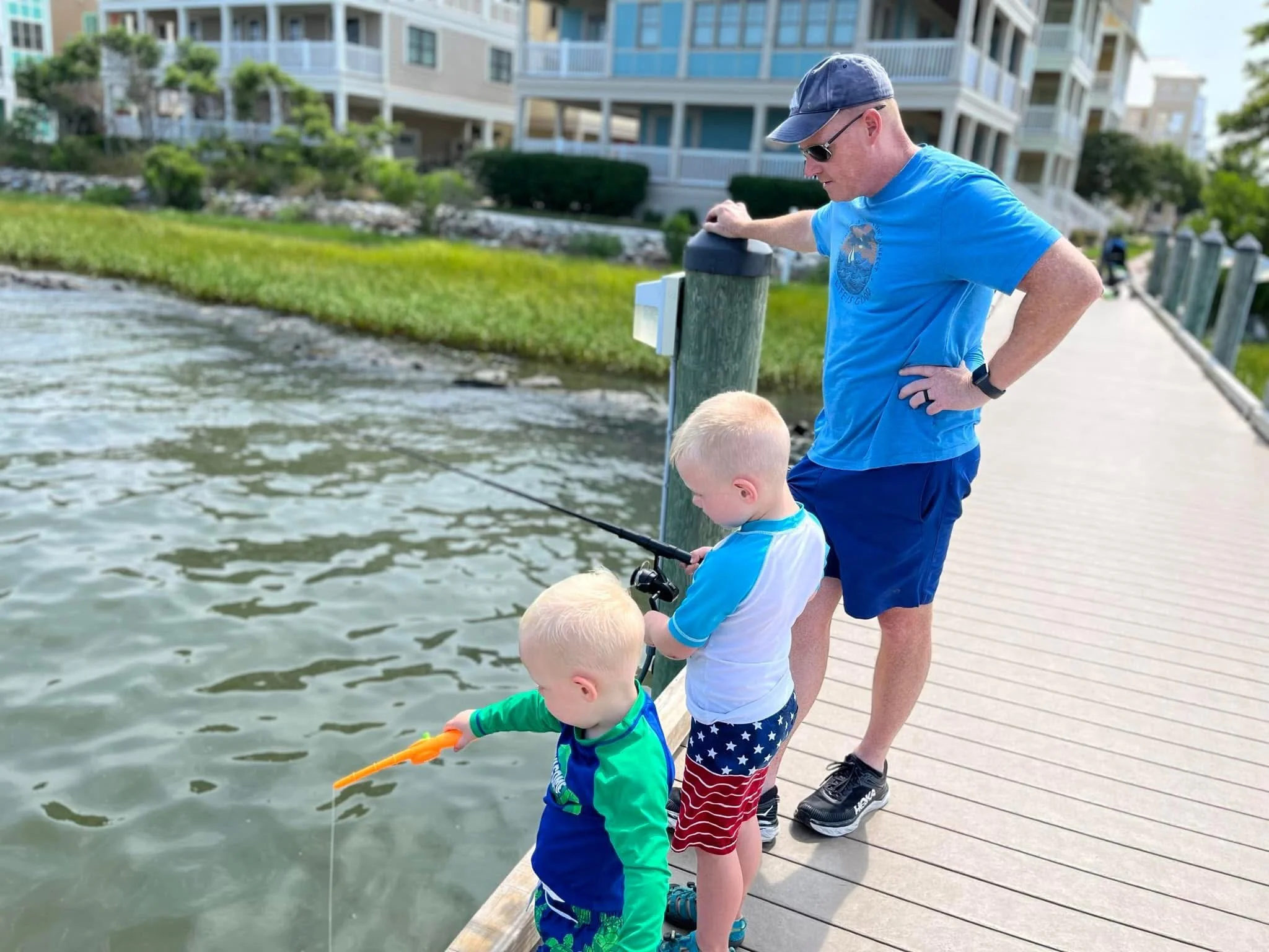 A man and three children fishing off a wooden dock by the water, with residential buildings and greenery in the background.