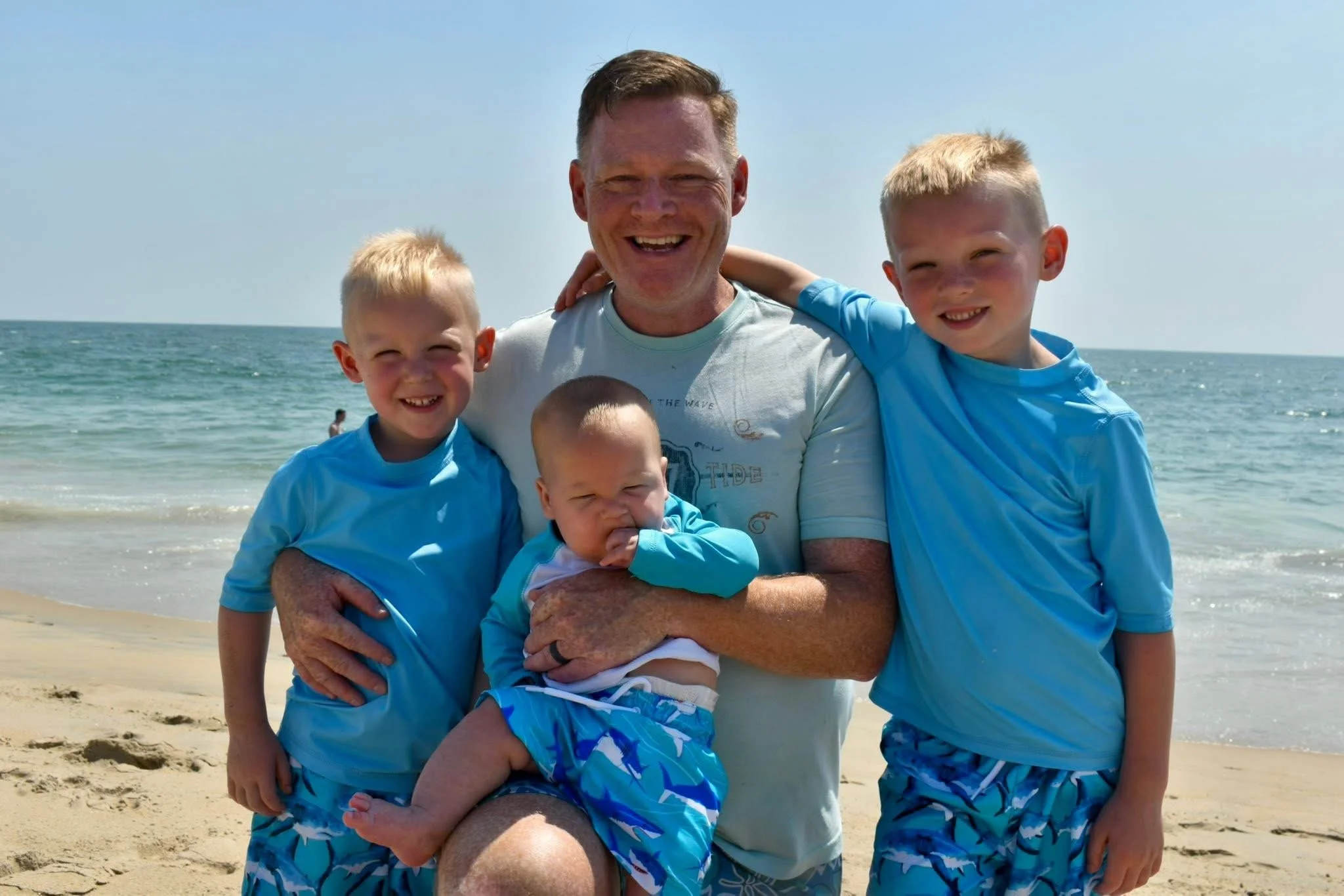 A man with four young boys at the beach, all wearing matching blue shirts and swim trunks, smiling and posing for the photo with ocean and clear sky in the background.