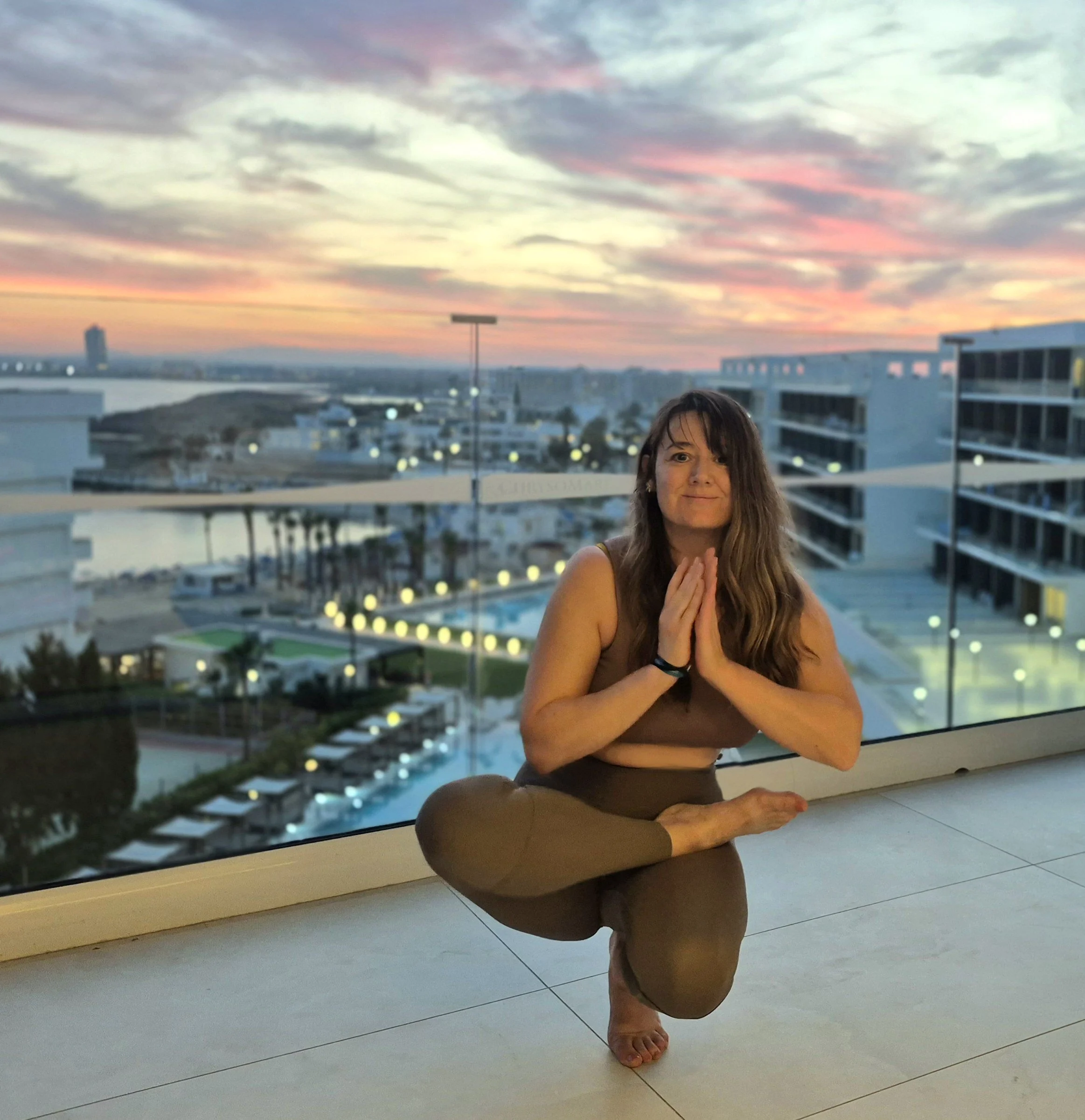 A woman practicing yoga on a balcony at sunset, mid pose with one leg bent and hands in prayer position, overlooking a cityscape with modern buildings, a pool, and a colorful sky.