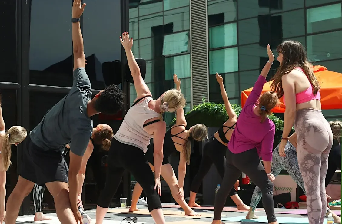 People participating in an outdoor yoga class, stretching on mats in front of a modern glass building.