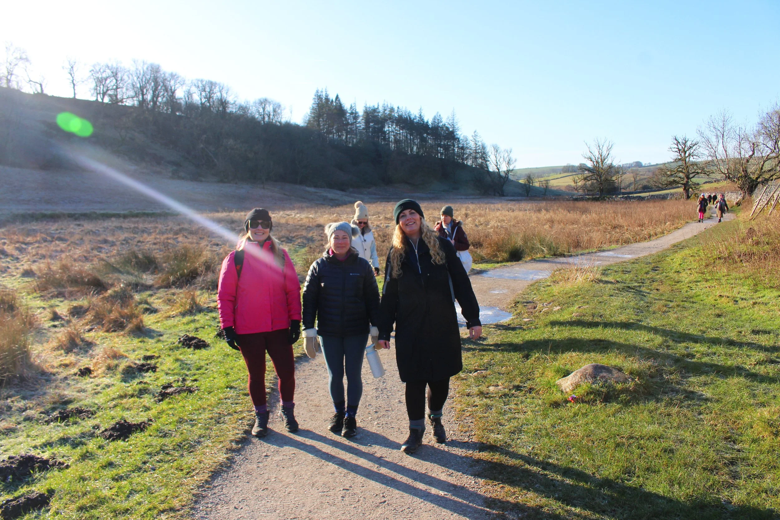 A group of people hiking outdoors on a bright, sunny day with clear skies, walking along a dirt trail through a grassy field with trees in the background.
