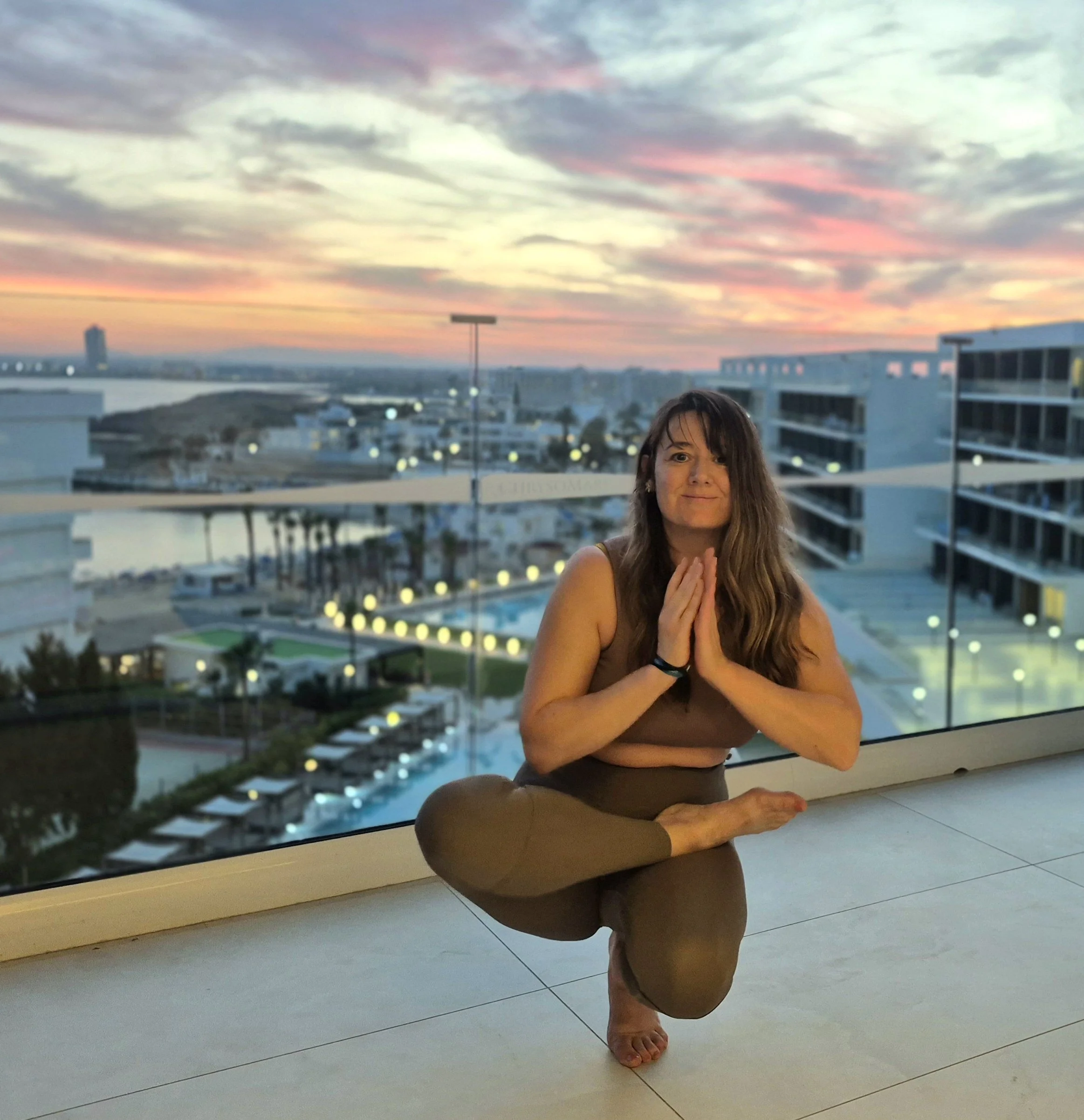 A woman practicing yoga on a balcony during sunset, with city buildings, a pool, and a colorful sky in the background.