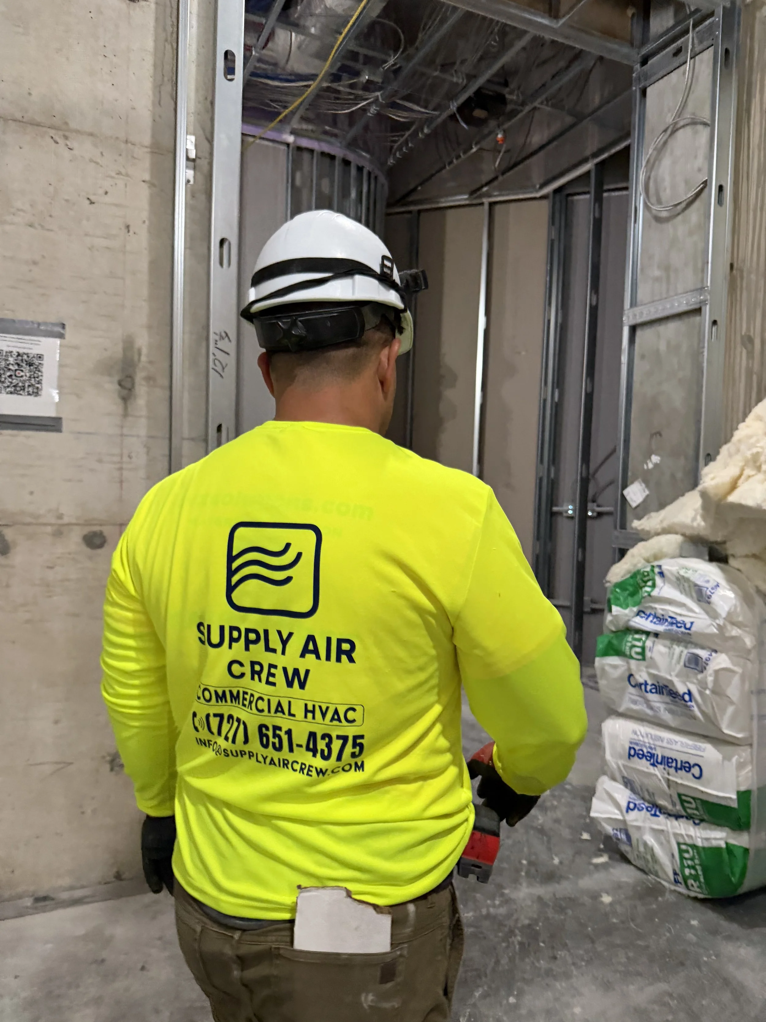 Construction worker wearing a yellow high-visibility shirt, a white safety helmet, and black gloves, standing inside a building under construction with metal framing and insulation materials nearby.