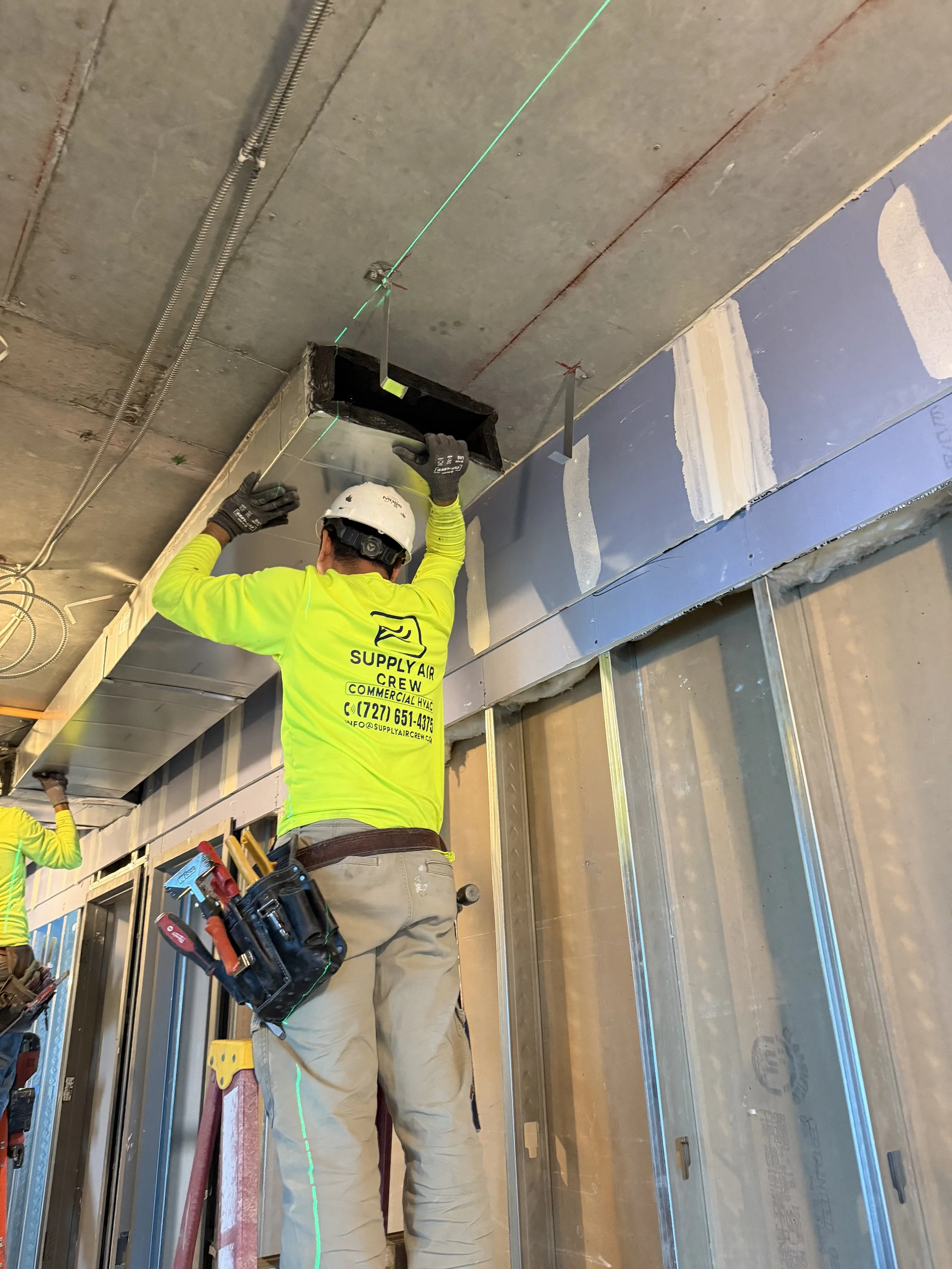Construction worker installing a black HVAC duct in a building under construction, using a green laser line for alignment, wearing a yellow shirt with company information and a safety helmet.