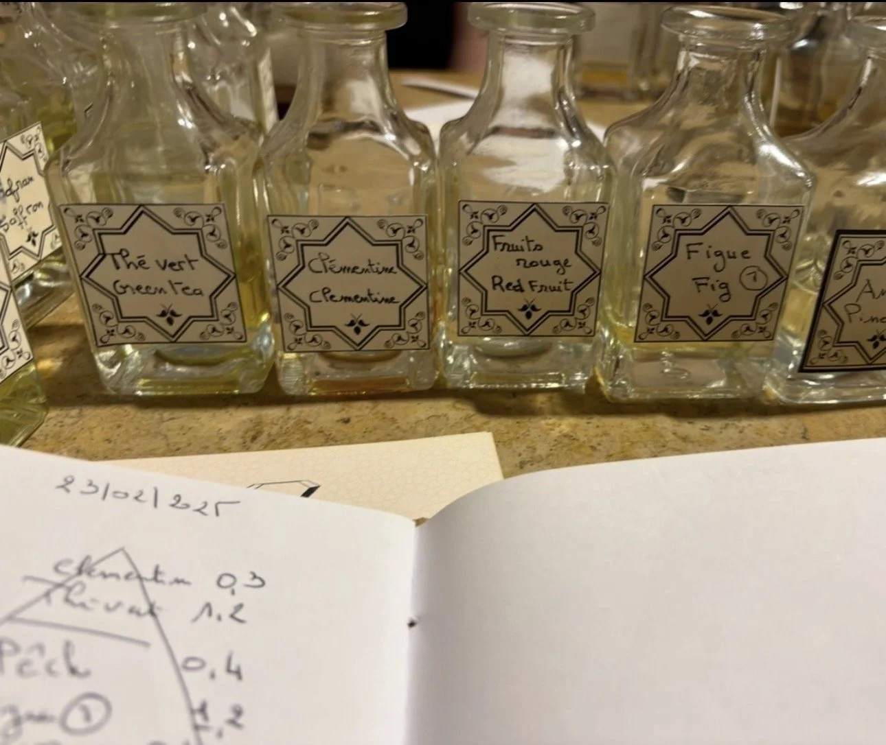 A row of glass bottles labeled with various fruit essential oils, including green tea, clementine, red fruit, and fig, placed on a countertop.