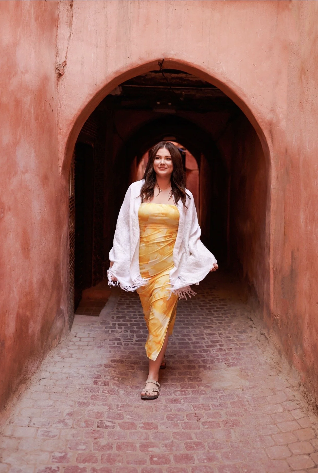 Woman in yellow dress and white shawl walking through an arched, peach-colored corridor with brick floor.