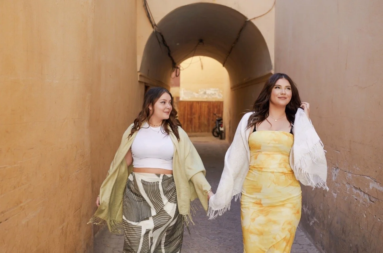 Two young women walking hand in hand through an alley with warm-colored walls, one in a yellow dress and the other in striped pants and a white crop top, both smiling and enjoying each other’s company.